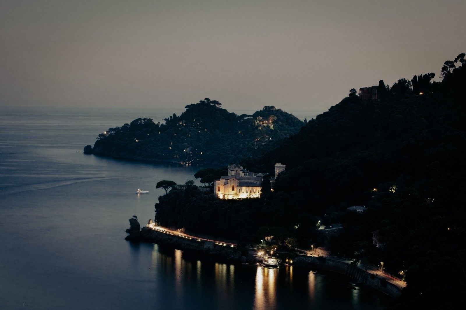 Twilight view of a serene coastline with illuminated buildings and lush hills overlooking calm waters.