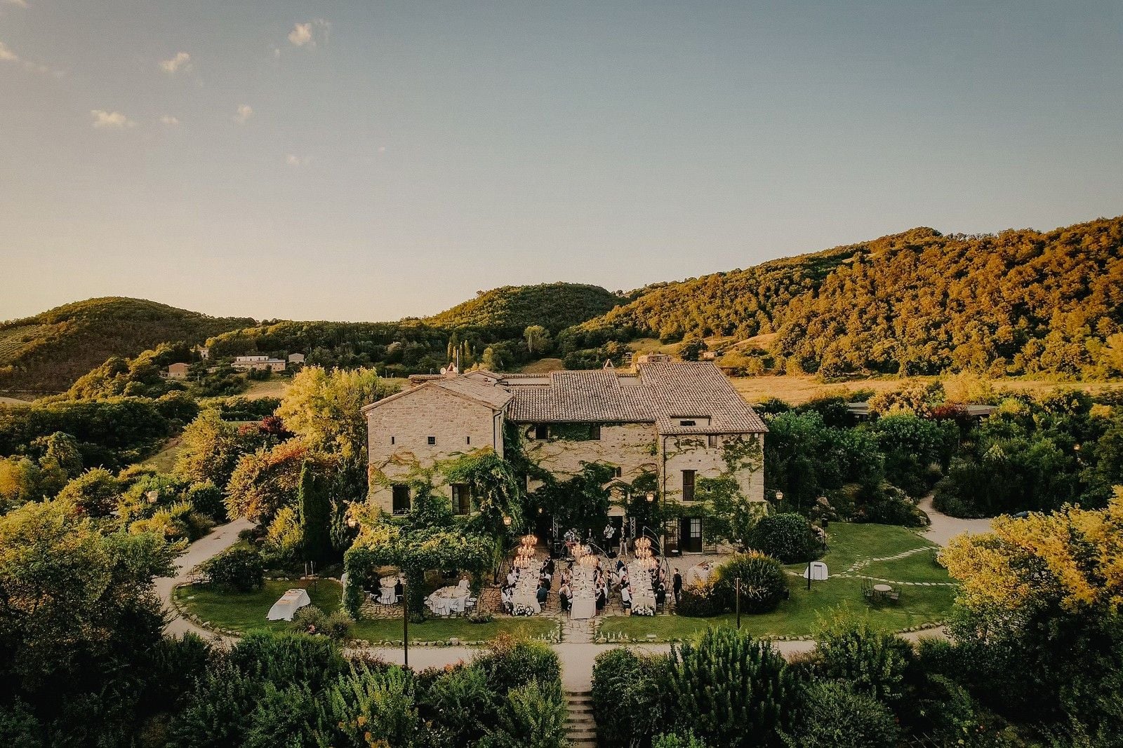A picturesque stone villa surrounded by lush greenery, hosting an outdoor event with guests mingling in the garden.