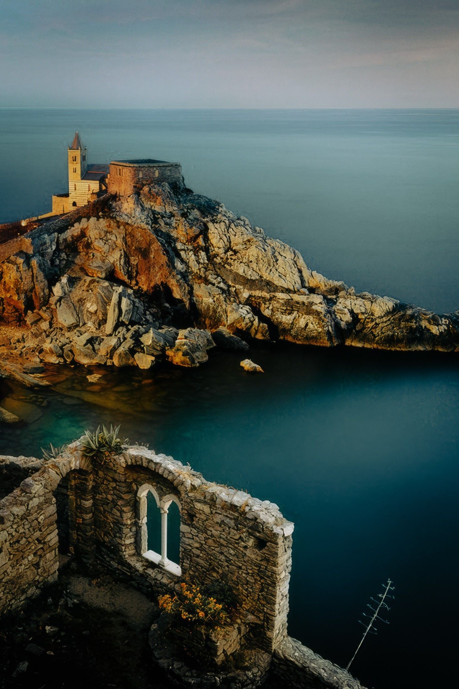 Historic stone structure atop a rocky island, with a lighthouse overlooking the calm sea at dusk.