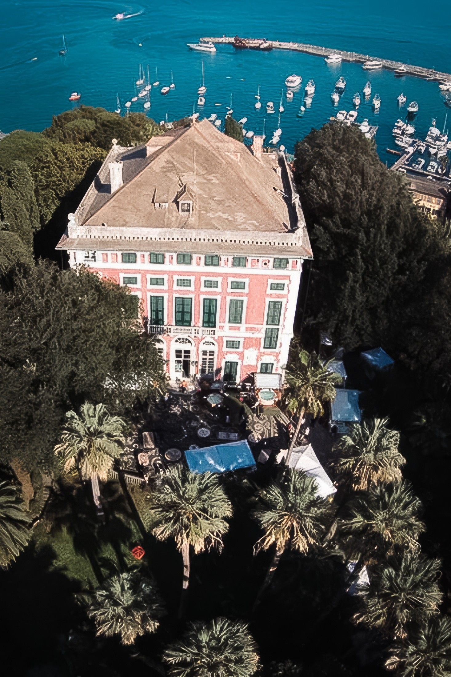 Historic villa overlooking a tranquil seaside harbor, surrounded by lush palm trees and sailboats in the distance.
