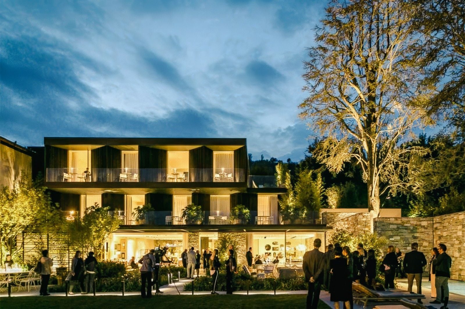 Elegant evening gathering outside a modern building surrounded by greenery, with guests socializing under a twilight sky.