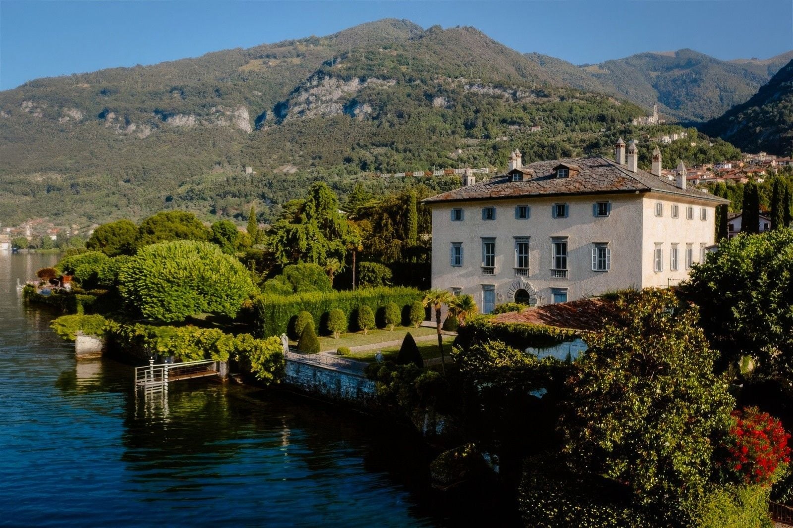 Historic villa situated by a tranquil lake, surrounded by lush greenery and mountains in the background.