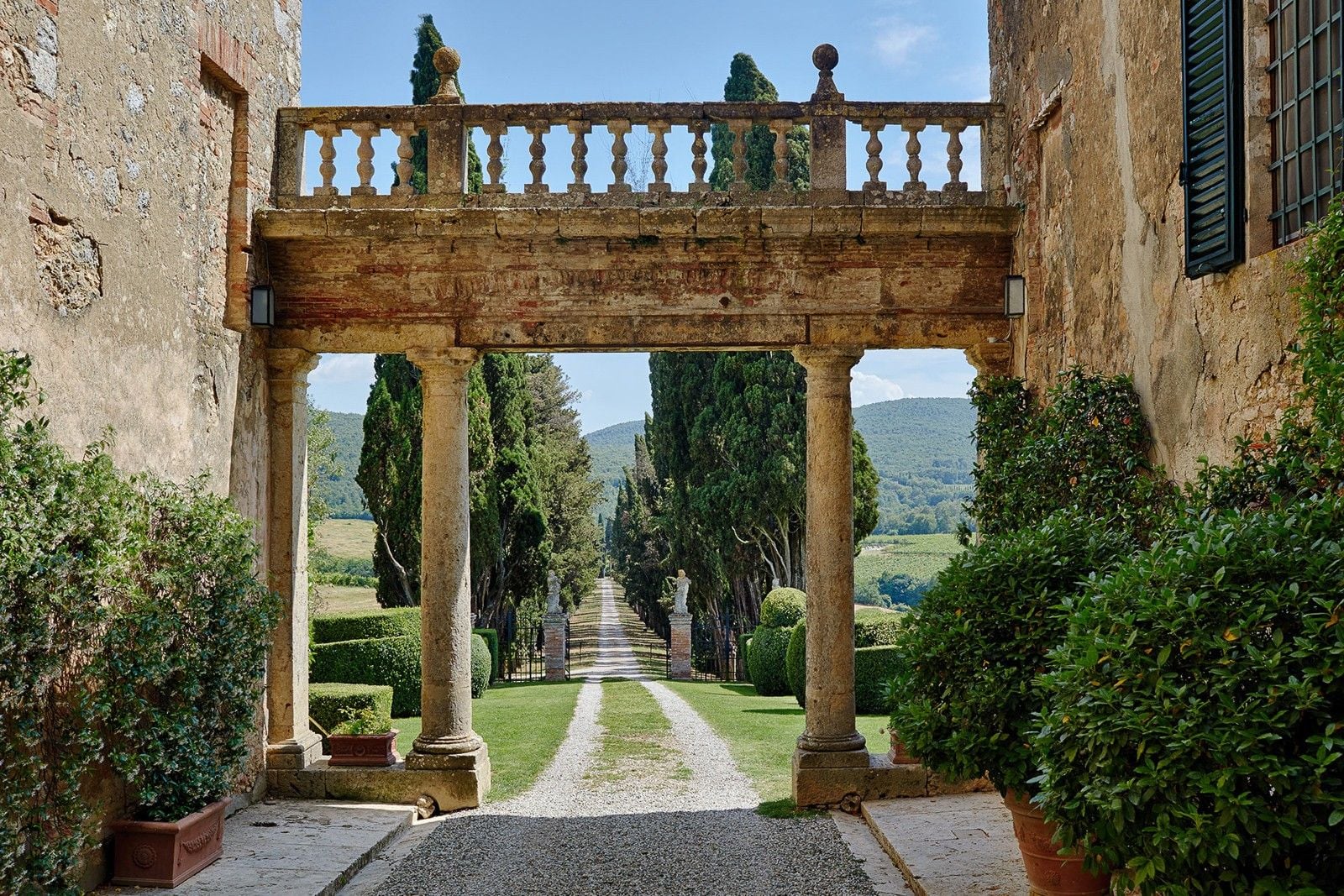 Historic stone archway leading to a lush garden pathway lined with cypress trees and ornamental hedges.