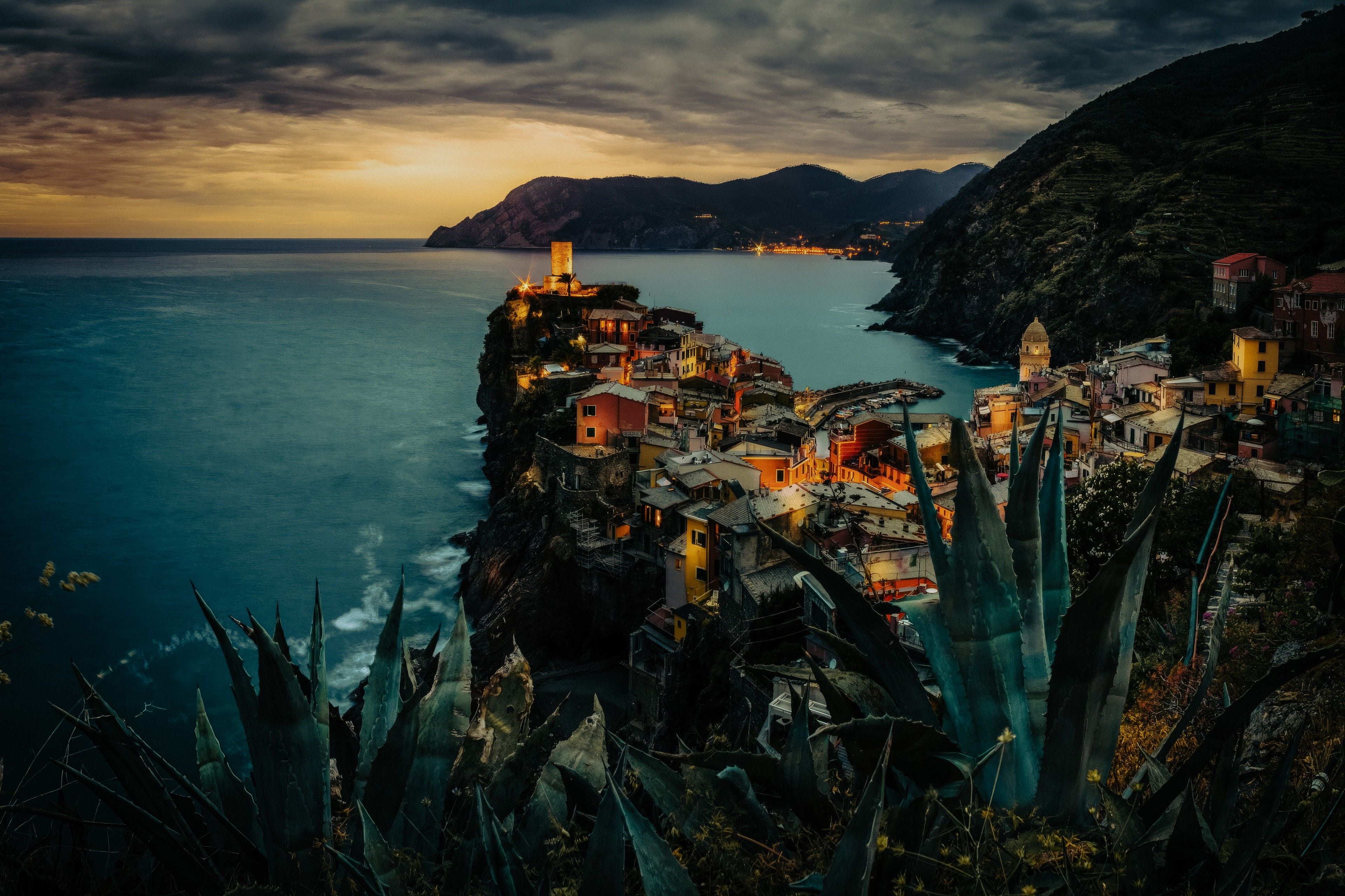 Colorful cliffside village of Vernazza twinkling at dusk, overlooking the serene sea with dramatic mountains in the background.