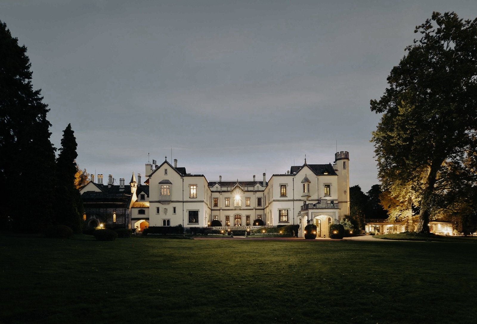 Historic mansion illuminated at dusk, surrounded by lush greenery and elegant landscaping.