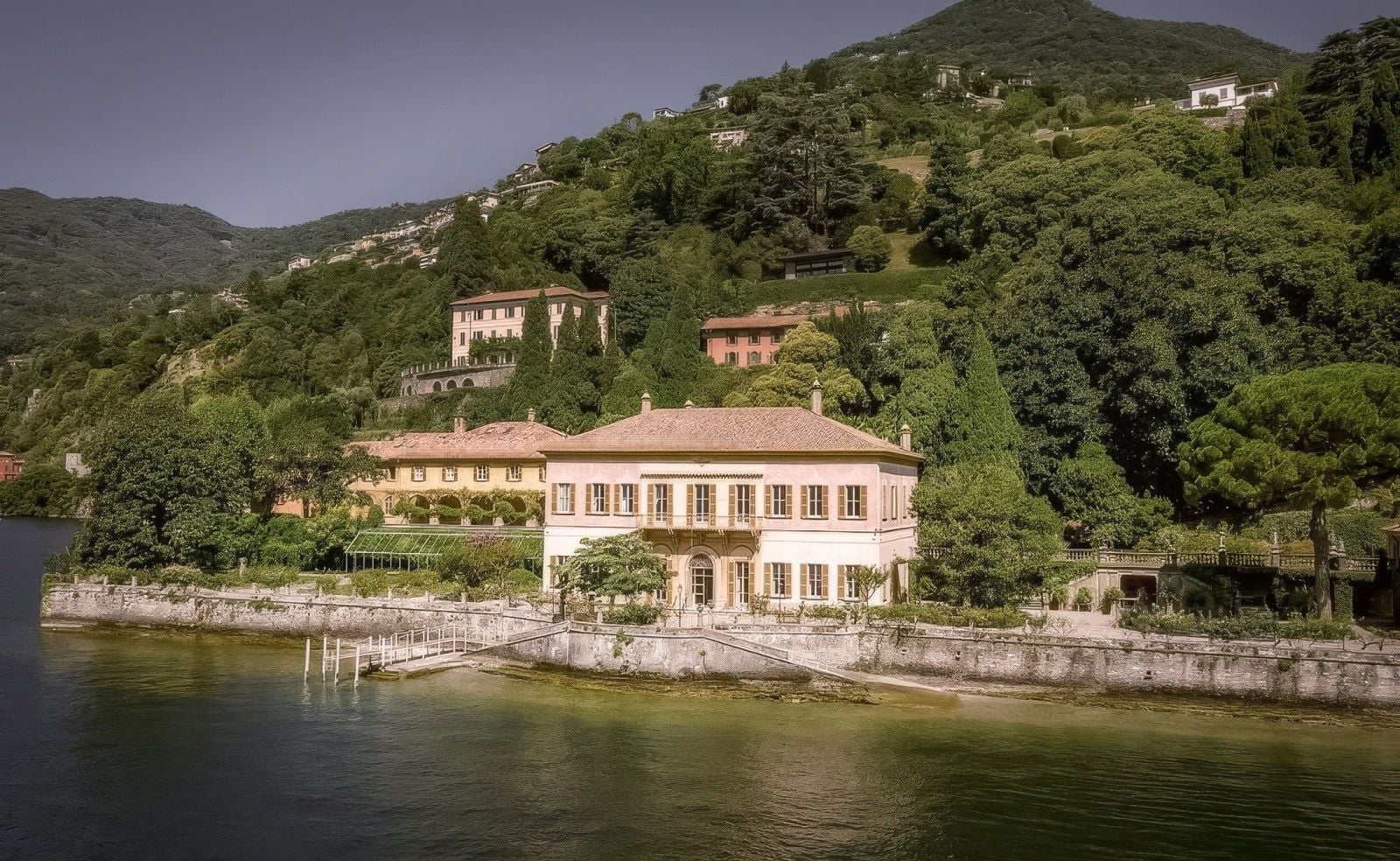 Elegant villa nestled by a serene lake, surrounded by lush greenery and hillside homes.