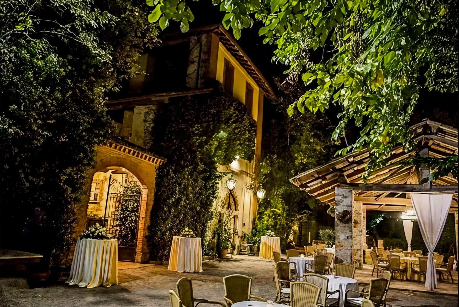 Elegant outdoor event setup surrounded by greenery, featuring tables and a pavilion at night.