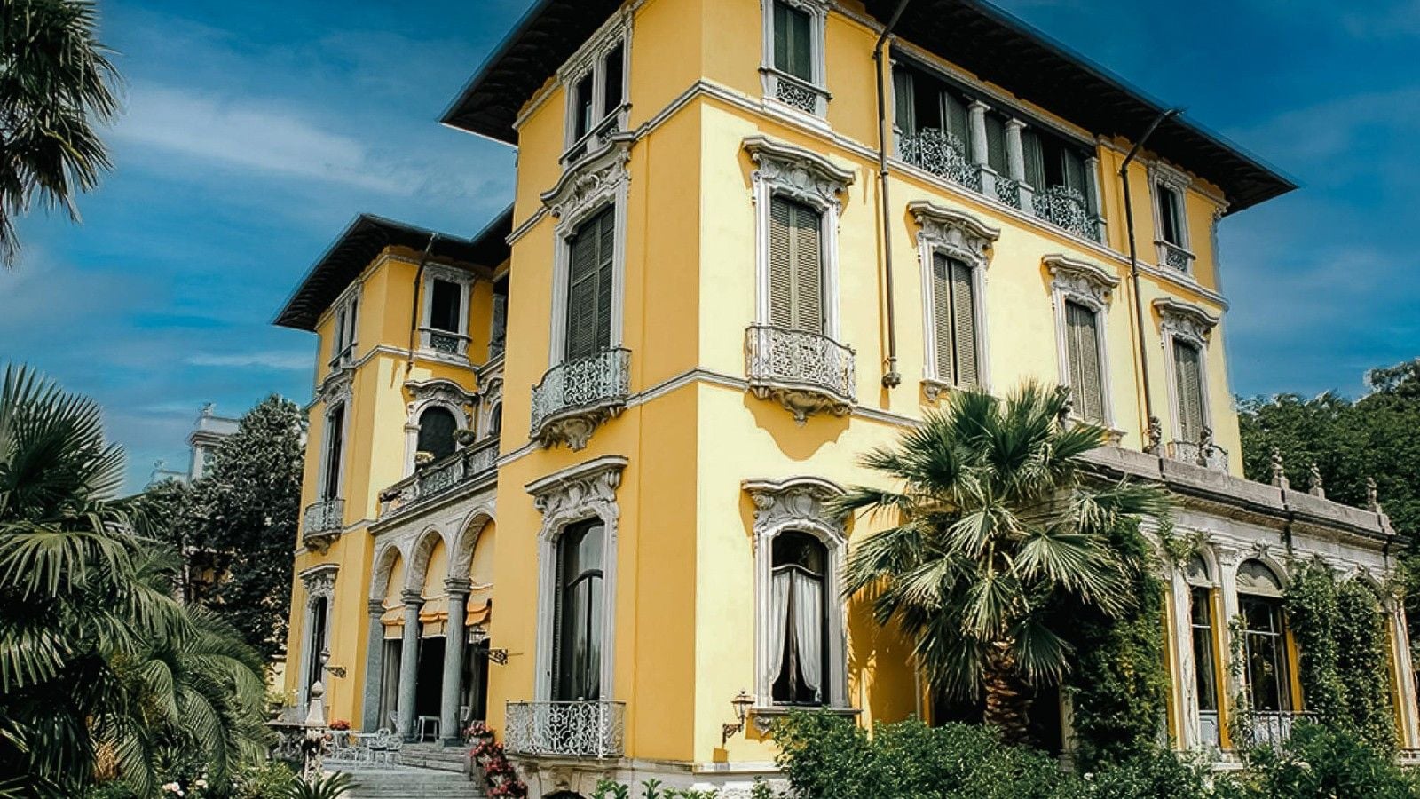 Elegant yellow villa with intricate balconies surrounded by lush greenery and palm trees under a blue sky.