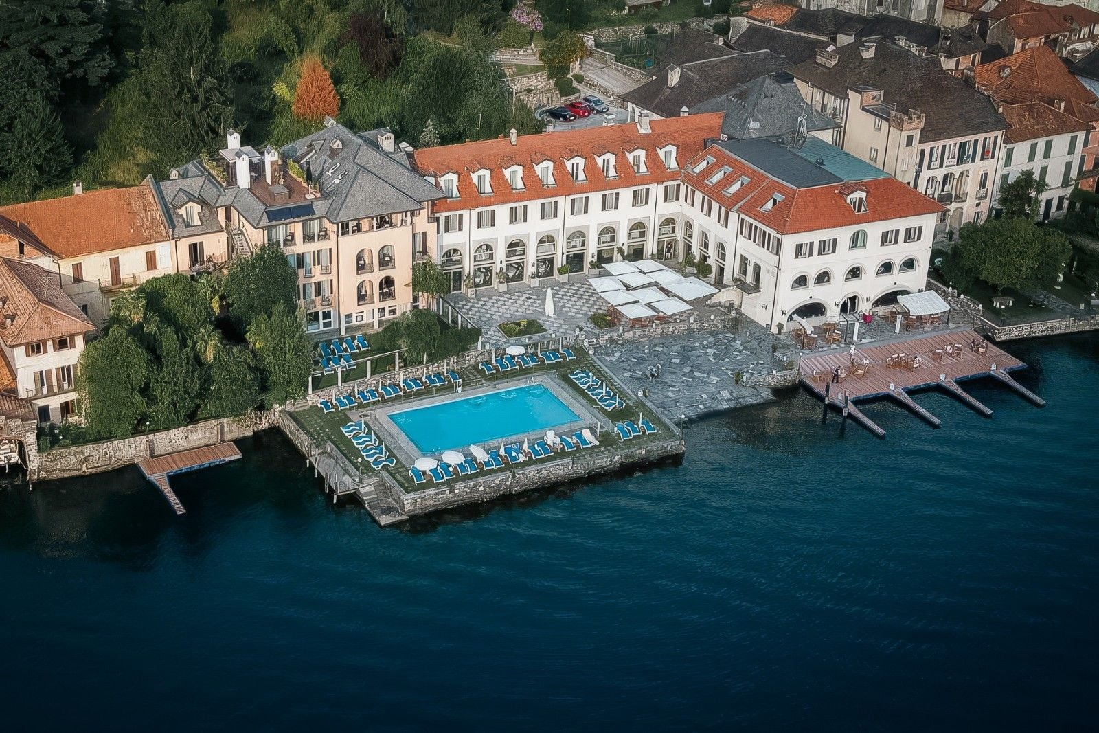 Luxury hotel by the lake featuring a pool, sun loungers, and elegant architecture surrounded by lush greenery.