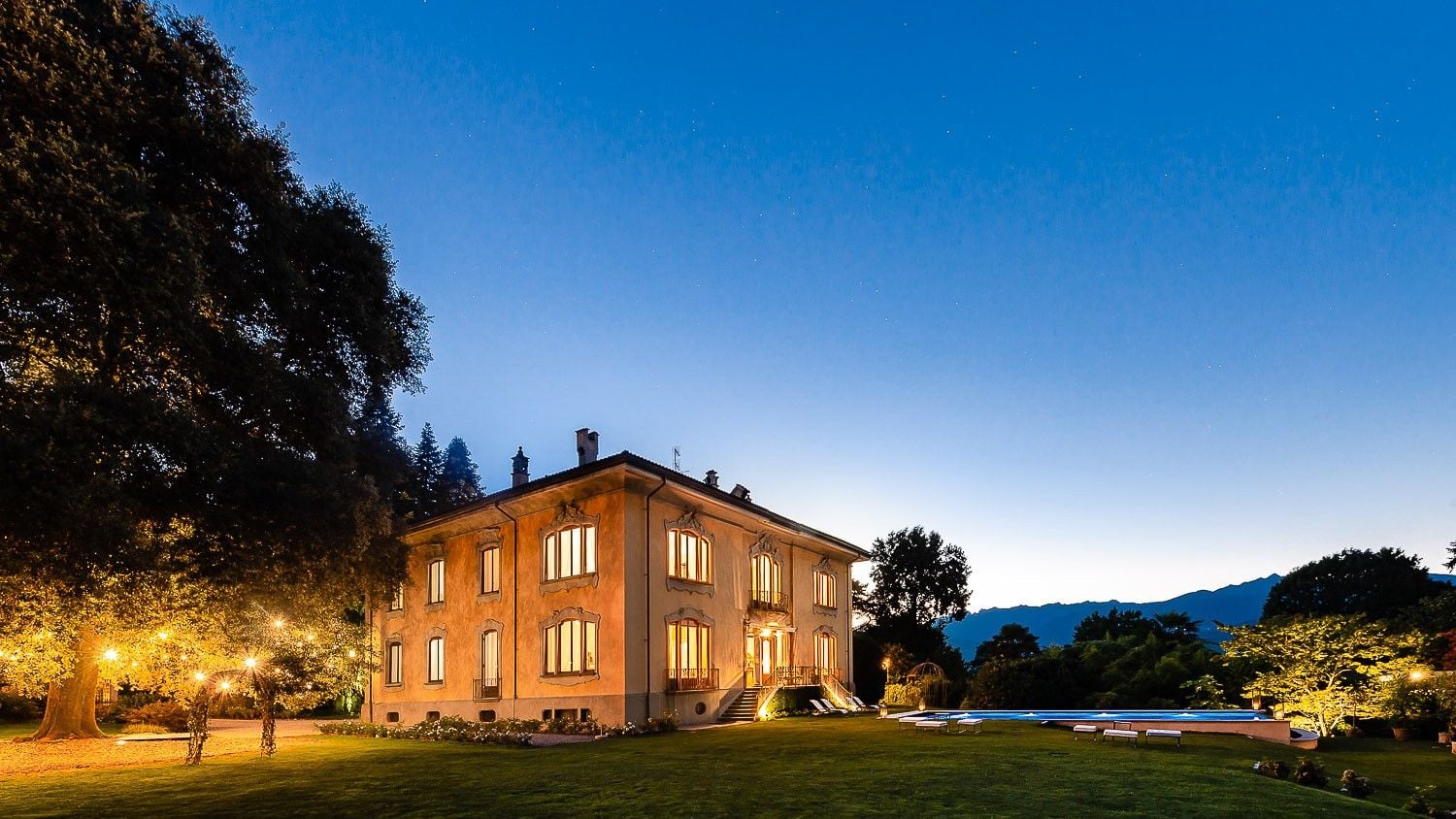 Elegant villa illuminated at dusk, surrounded by lush greenery and a serene pool under a twilight sky.