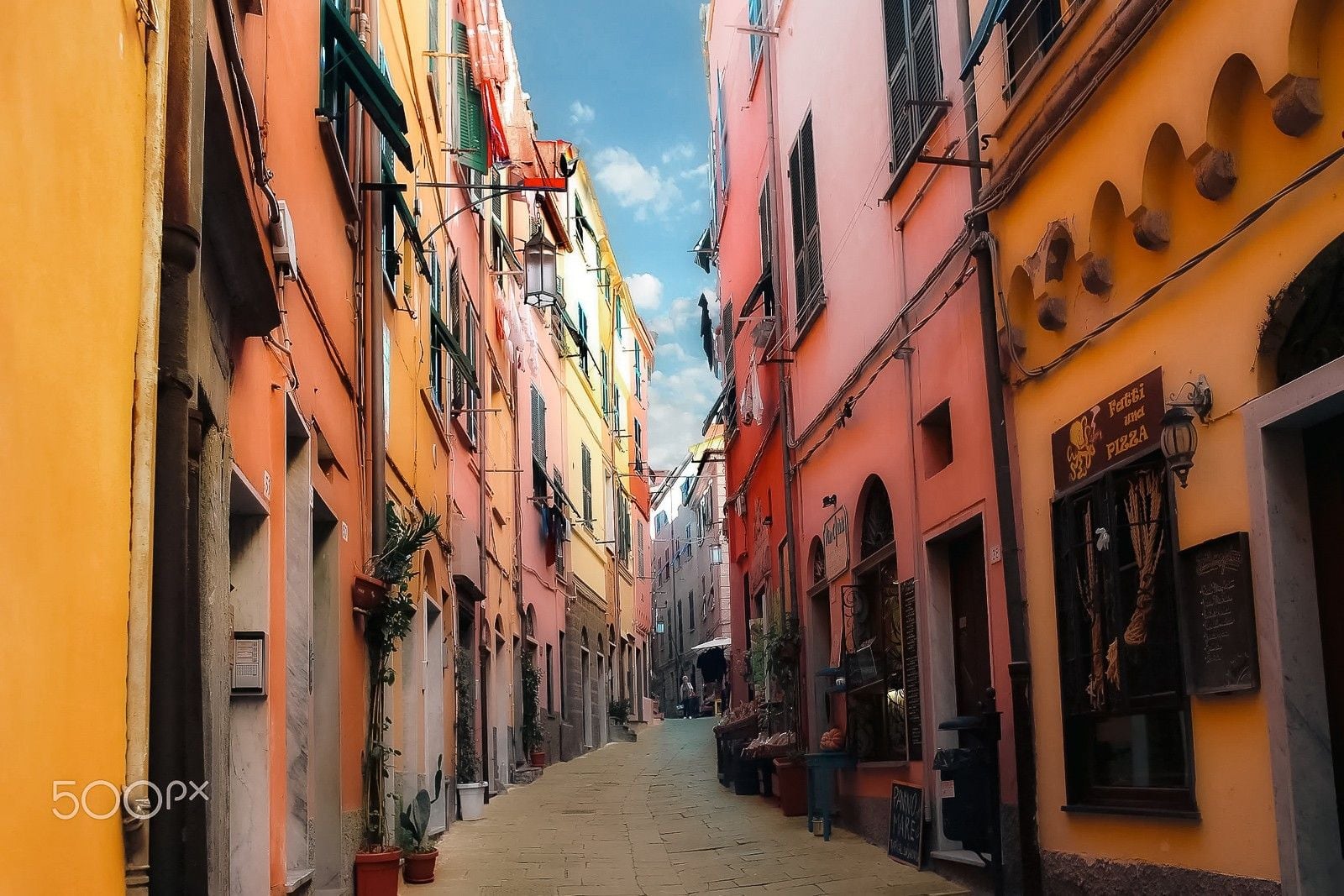 Narrow cobblestone street lined with colorful buildings, café signs, and vibrant flower pots in a charming Italian village.
