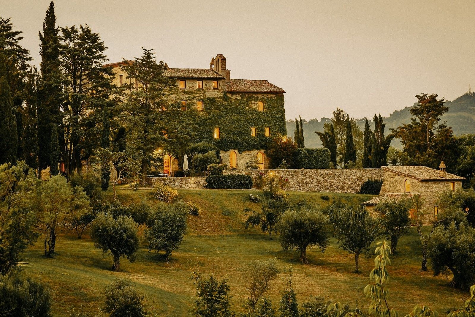 A rustic villa surrounded by lush greenery and trees, illuminated softly in the evening light.
