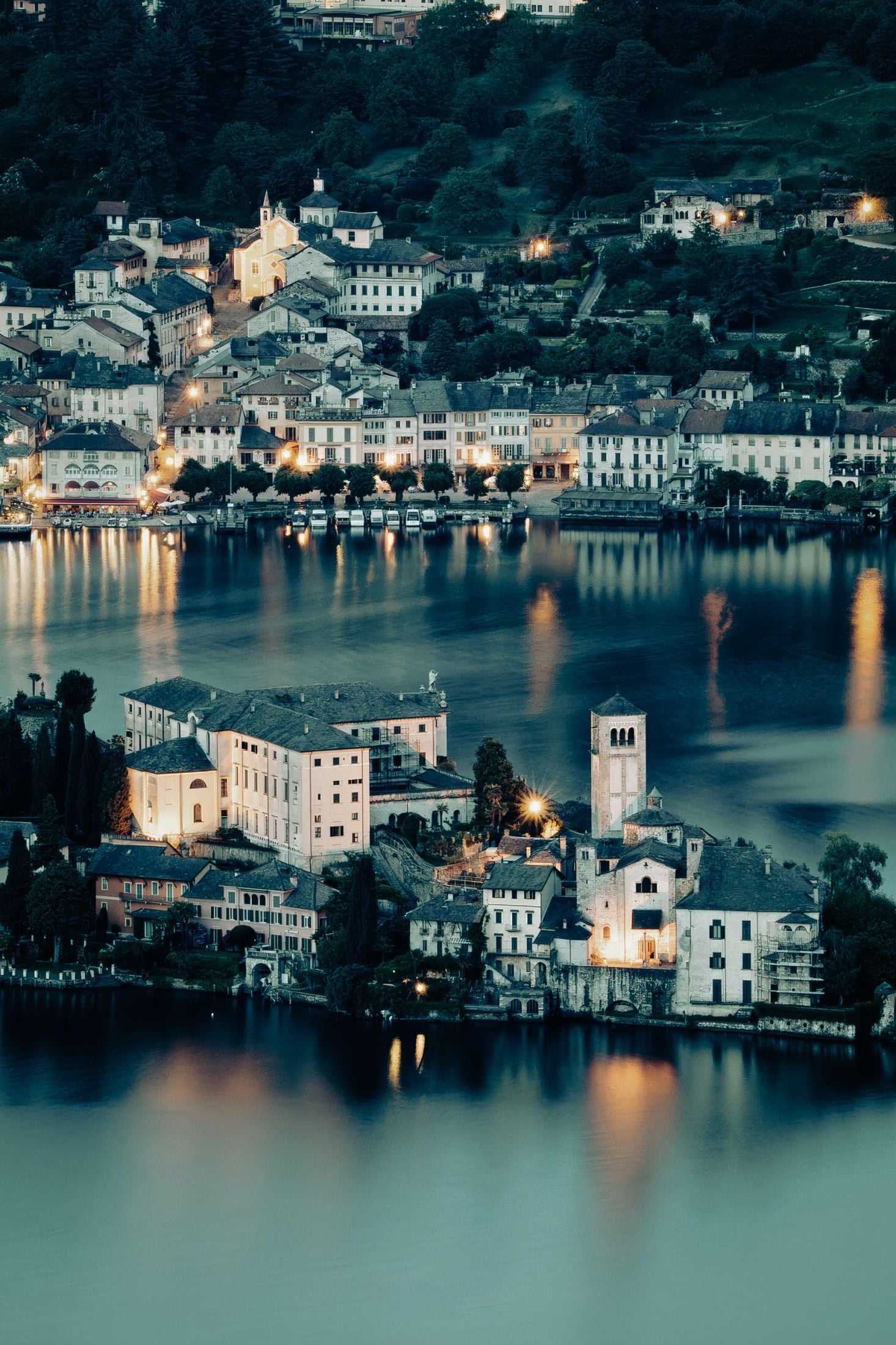 Picturesque lakeside village at twilight, reflecting lights on calm water, surrounded by serene hills and lush greenery.