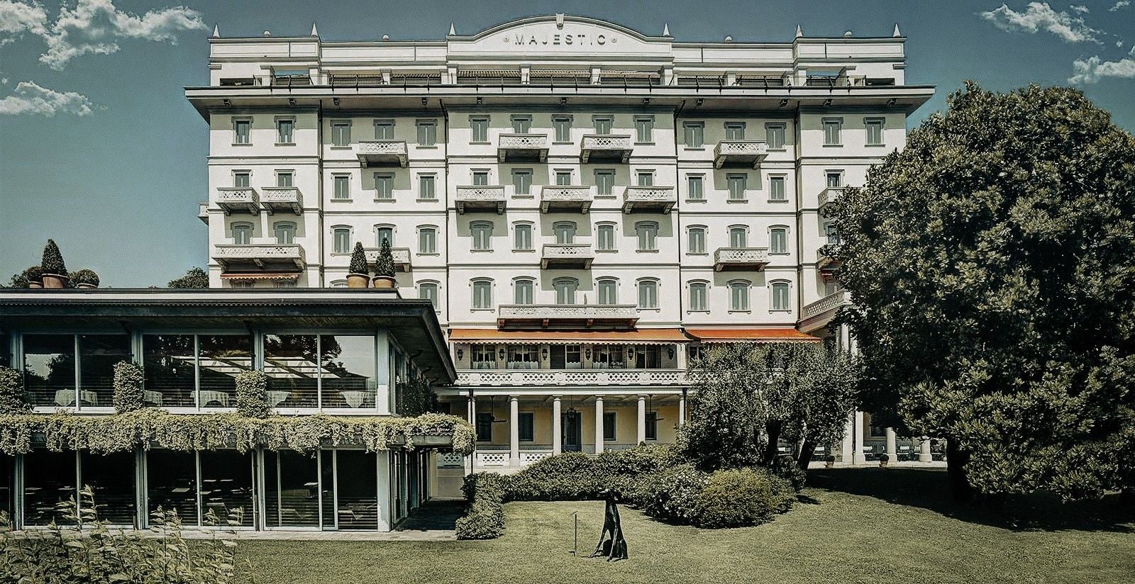 Elegant historic hotel facade surrounded by lush greenery, with a modern glass extension and clear blue sky.