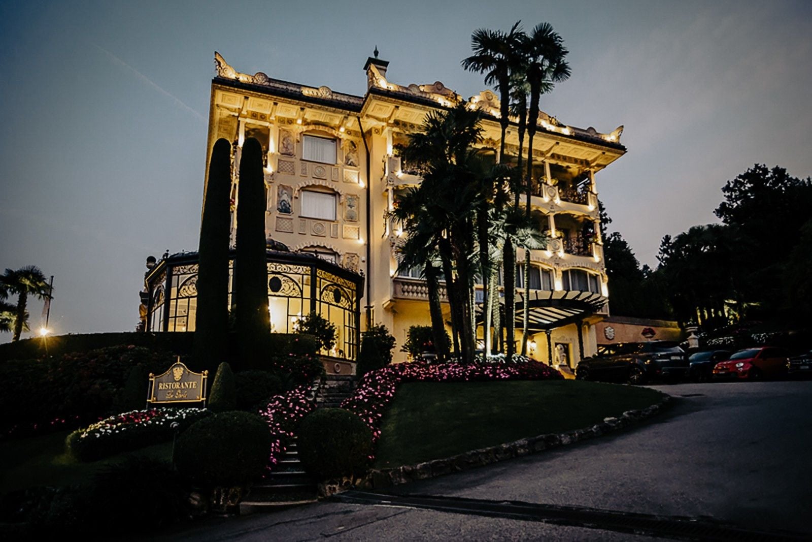 Luxurious mansion illuminated at dusk, surrounded by lush gardens and decorative plants.