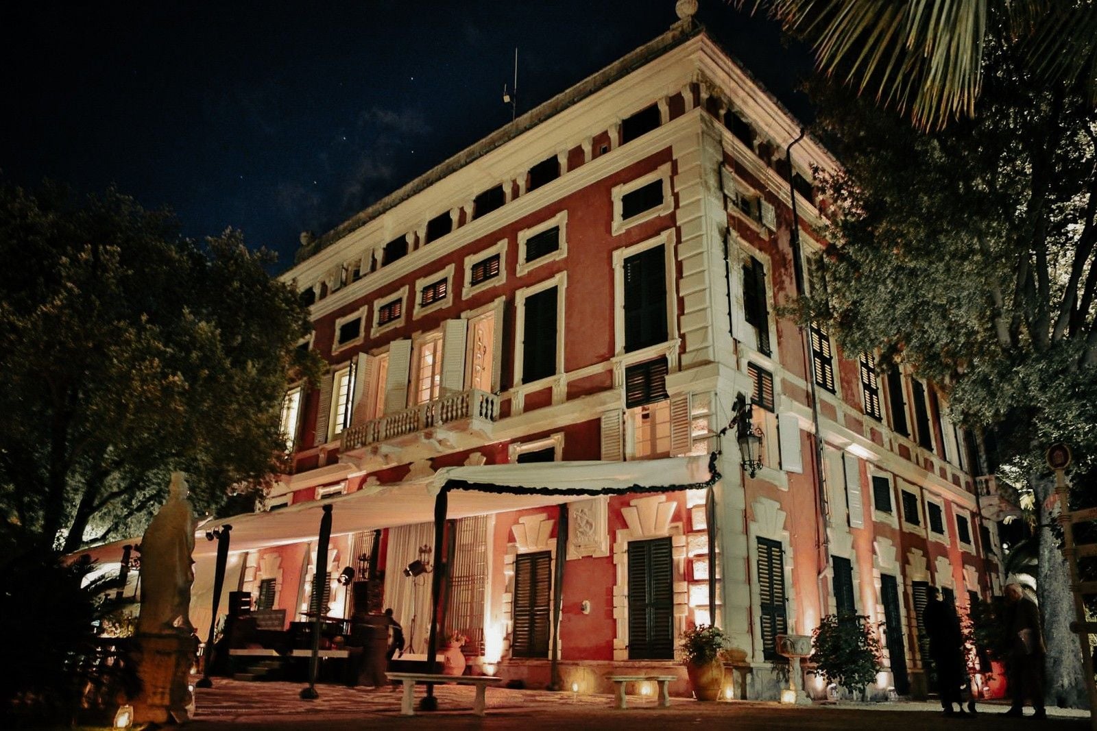 Elegant historic building with red and white façade, warmly lit at night, surrounded by lush greenery.