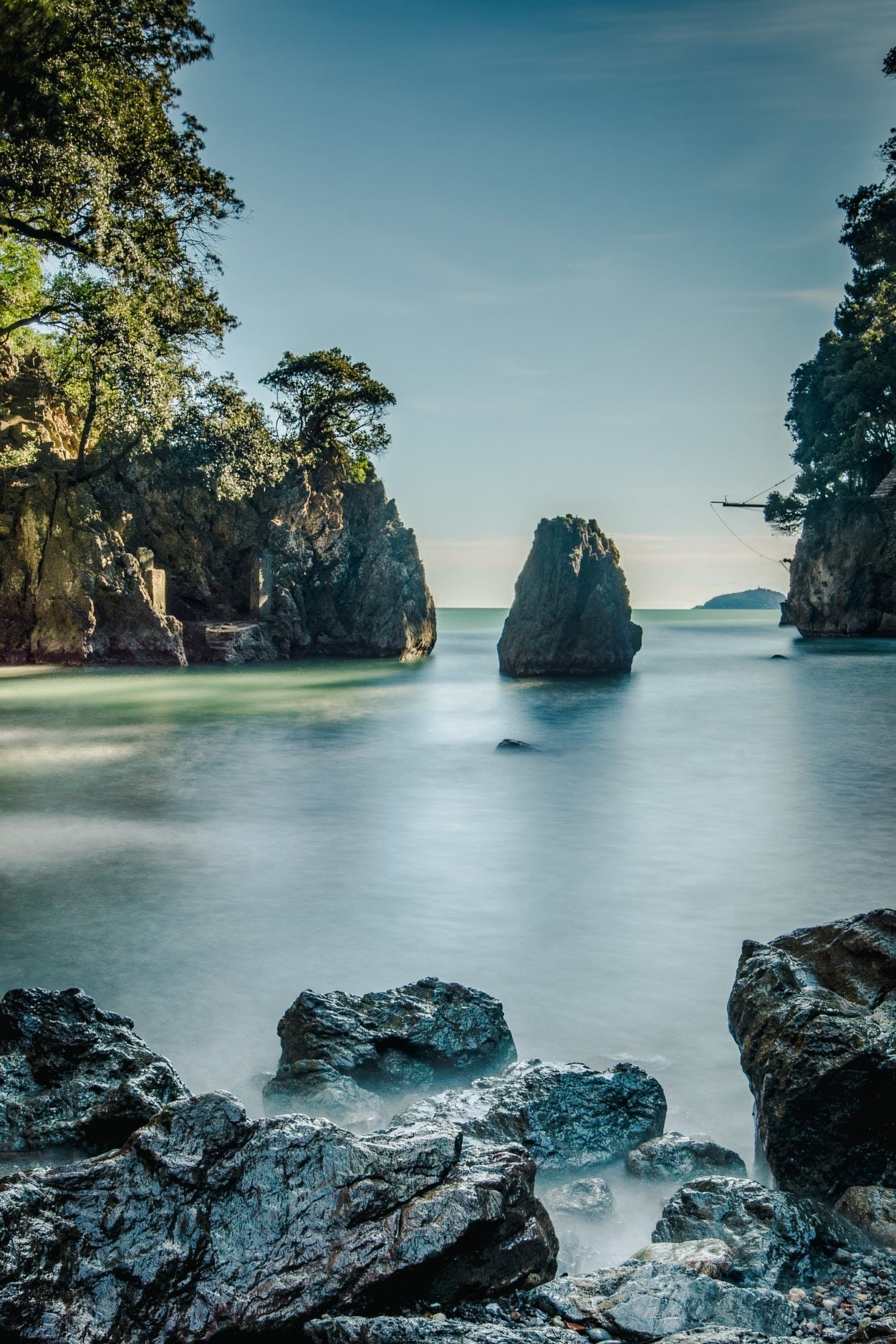 Serene coastal scene featuring smooth water, rocky formations, and lush greenery under a clear sky.