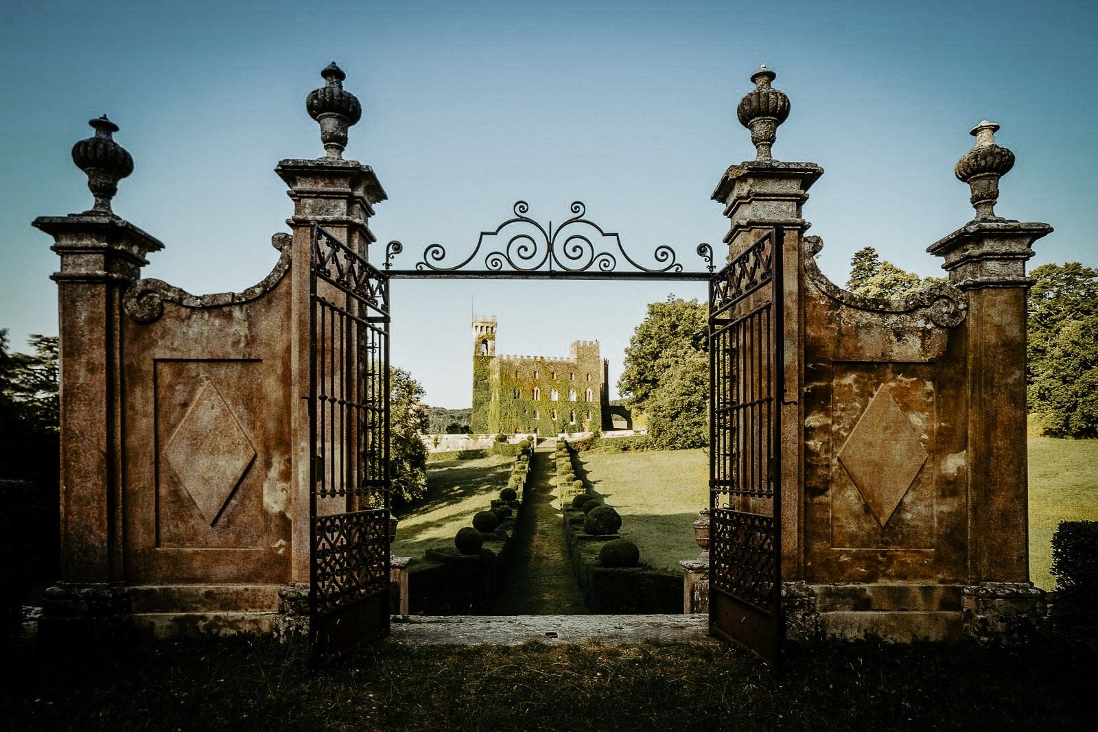 A grand iron gate opens to a lush garden path leading to a historic tower surrounded by greenery.