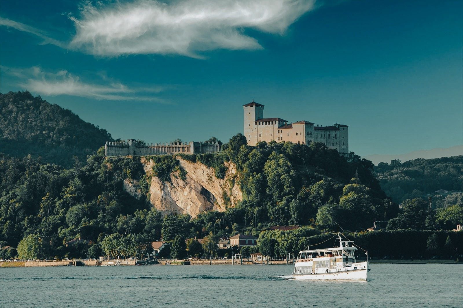 A sleek white boat sails past a historic castle perched on a rocky hill, surrounded by lush greenery and a serene sky.
