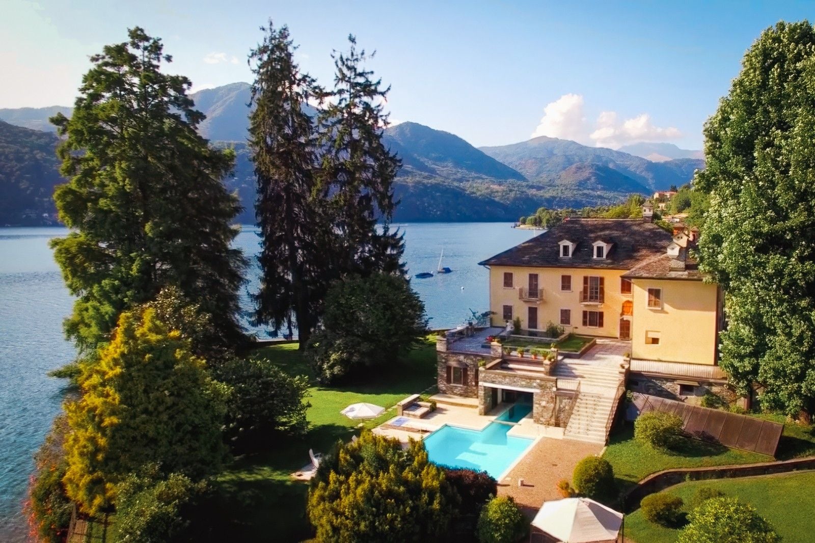 Scenic lakeside villa with a pool, surrounded by lush greenery and mountains under a clear blue sky.