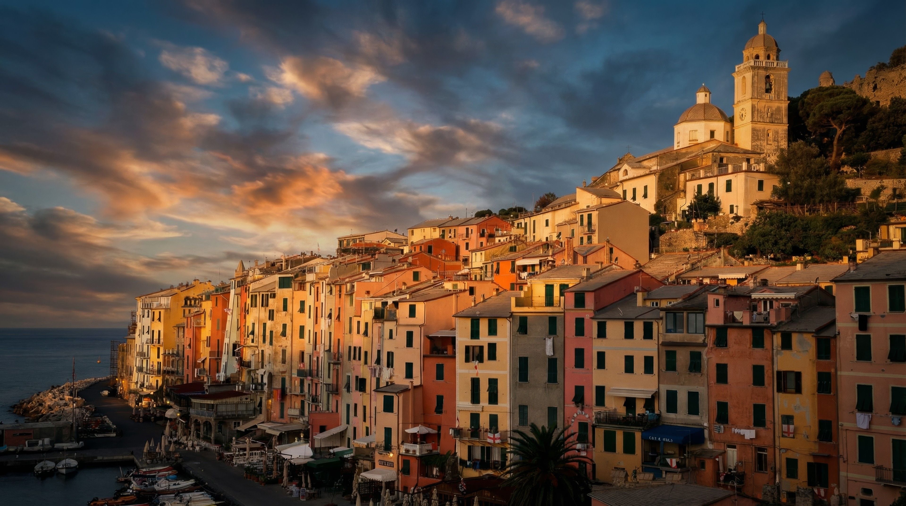 Sunset over a coastal village, showcasing colorful buildings nestled beside a serene harbor and dramatic sky.