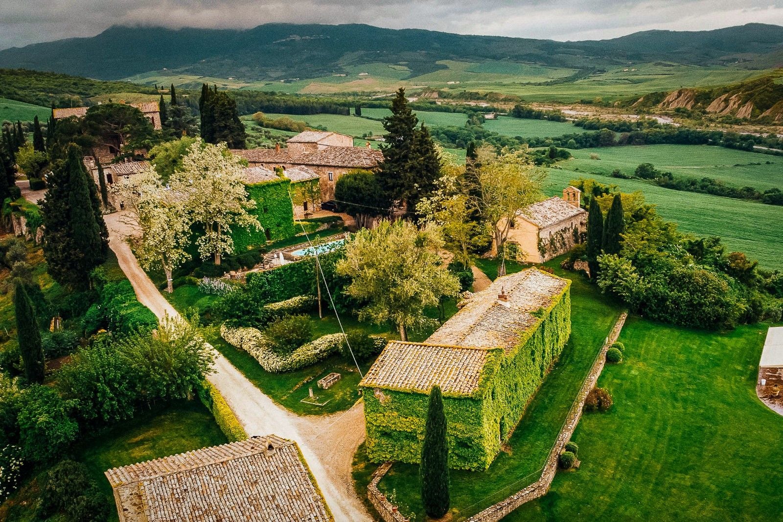 Aerial view of a lush Italian landscape featuring vine-covered buildings and rolling green hills under a cloudy sky.
