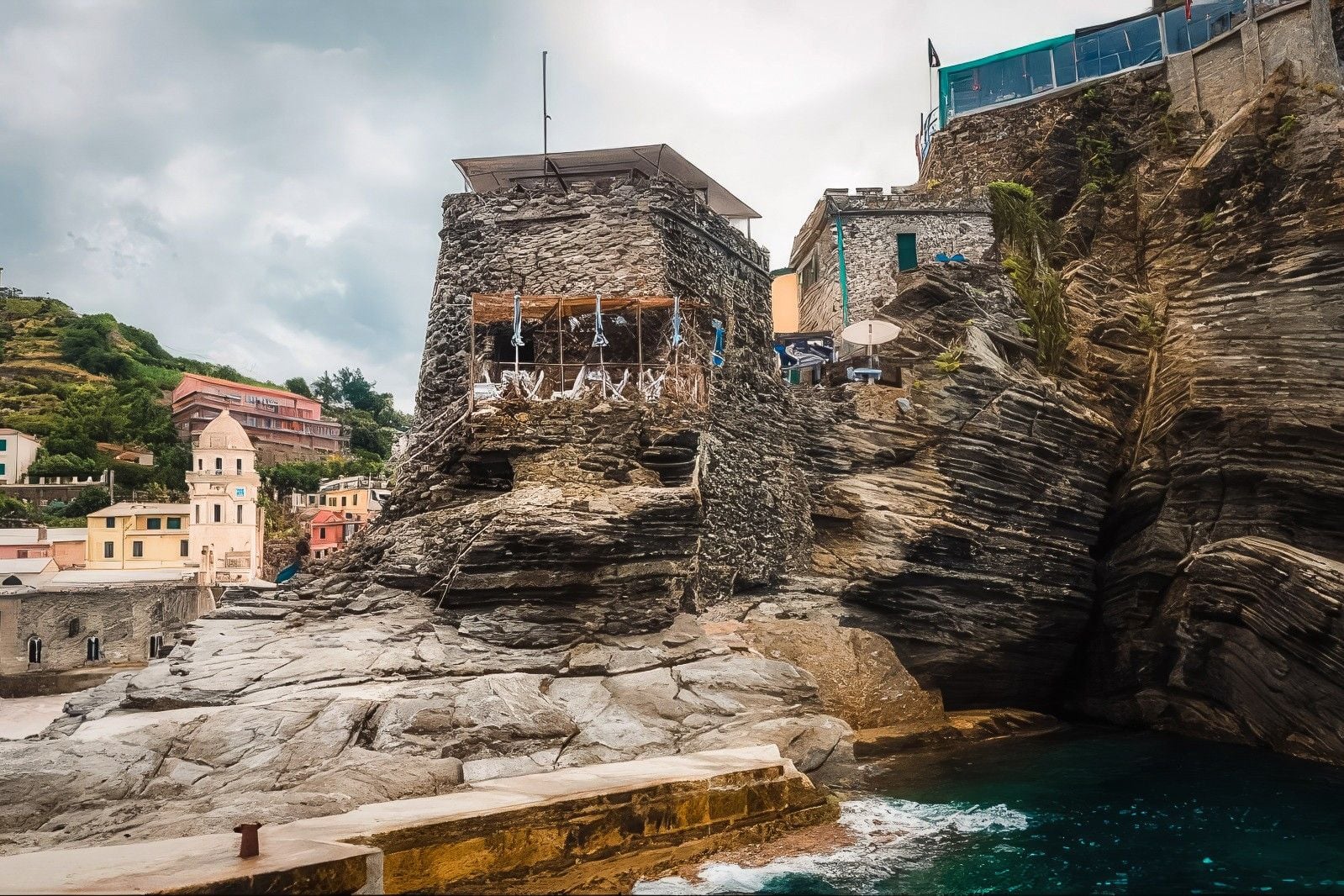 Unique rocky coastal structure overlooking tranquil turquoise waters, surrounded by colorful buildings and lush greenery.