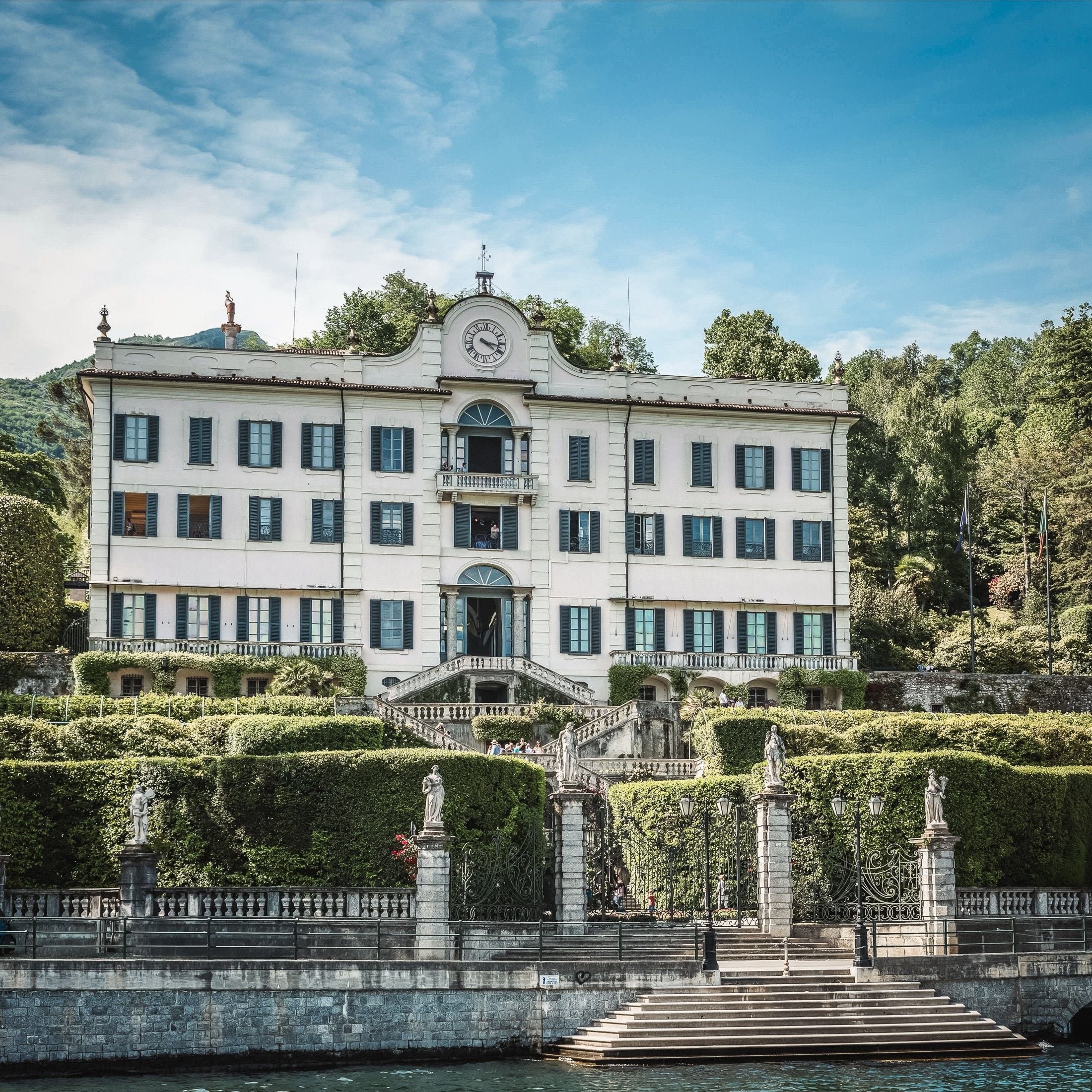 Elegant lakeside villa surrounded by lush gardens and ornate statues, under a clear blue sky.