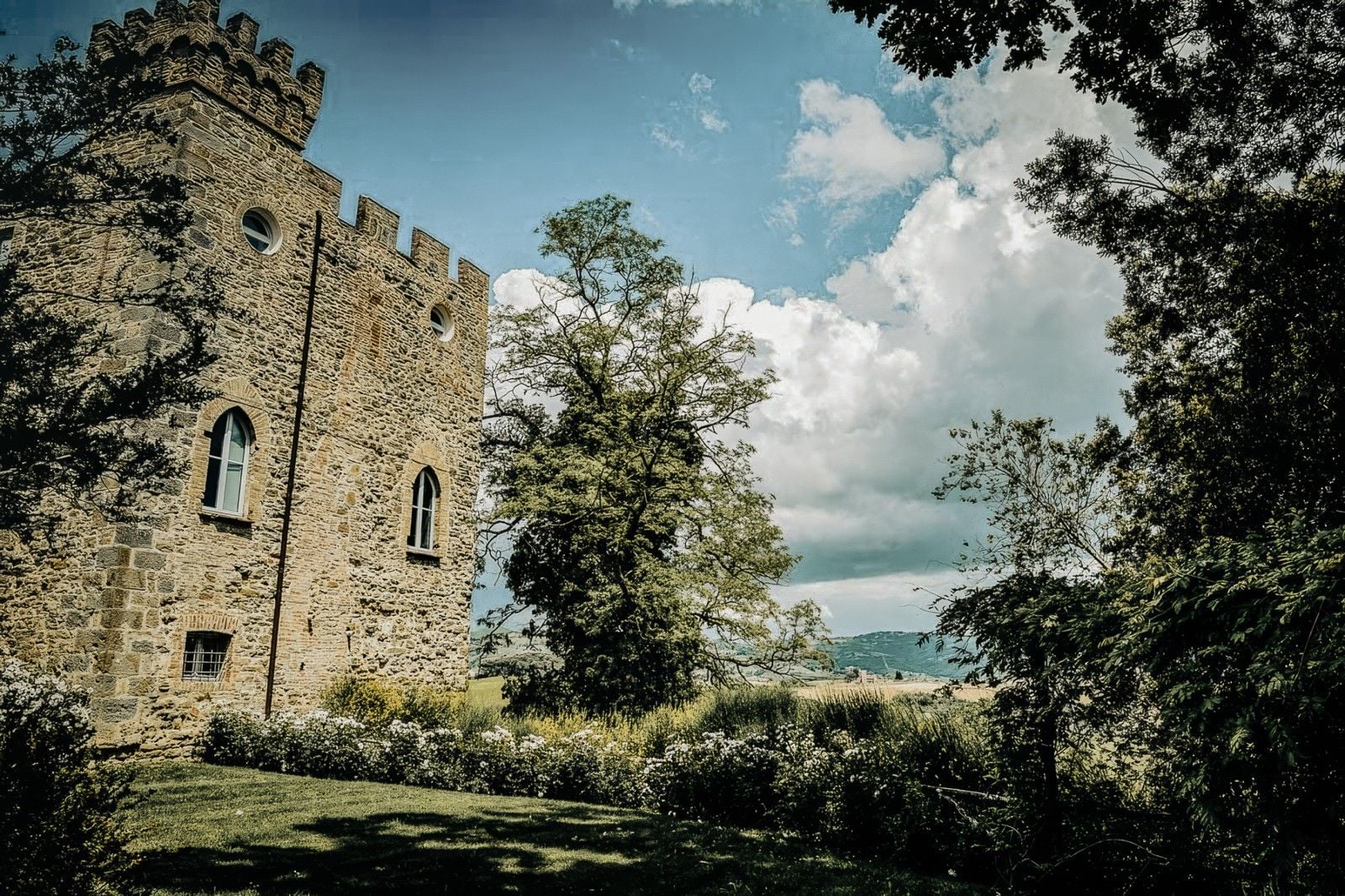 Historic stone tower surrounded by lush greenery and a scenic landscape under a partly cloudy sky.