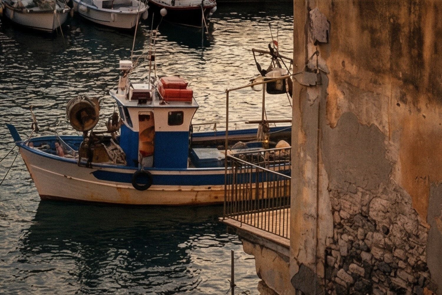 A small blue fishing boat moored near a weathered stone building, reflecting warm light on the water's surface.