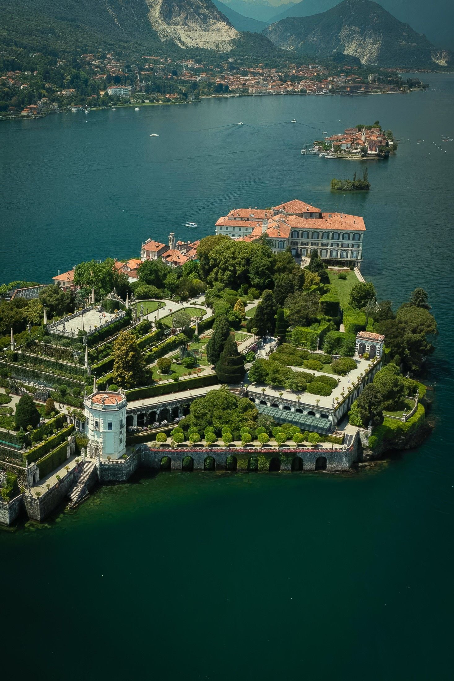 Aerial view of a grand villa surrounded by lush gardens and serene lake waters, with mountains in the distance.