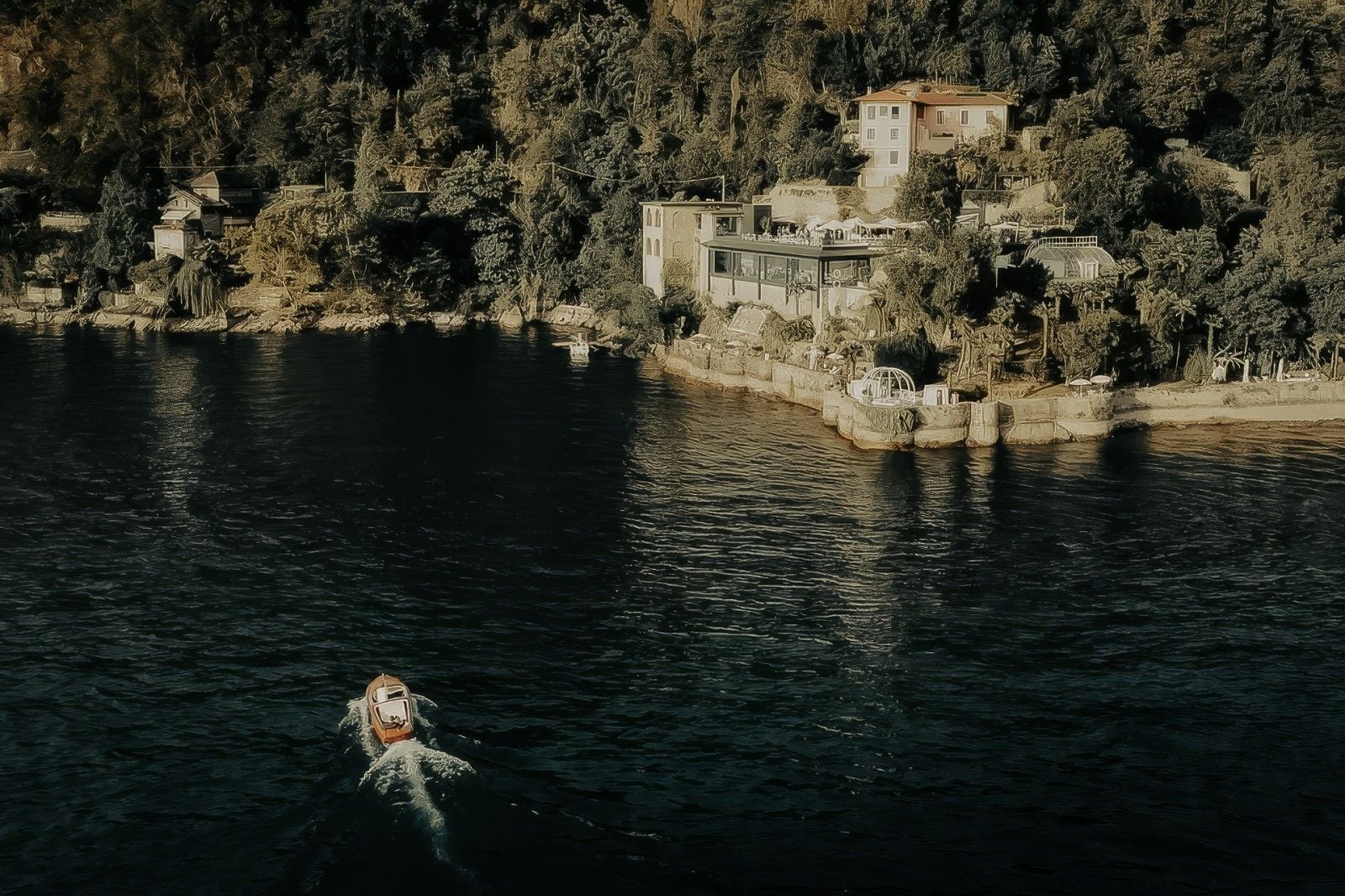 A small boat glides across calm waters near a lush, picturesque shoreline lined with elegant homes and greenery.