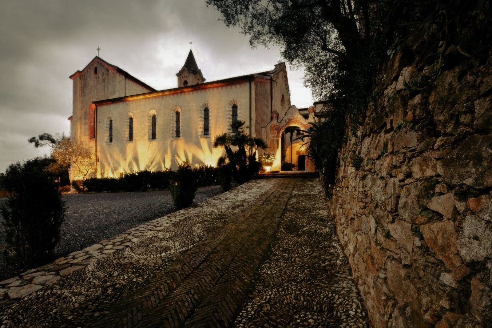 Historic church illuminated at dusk, surrounded by lush greenery and a textured stone pathway.