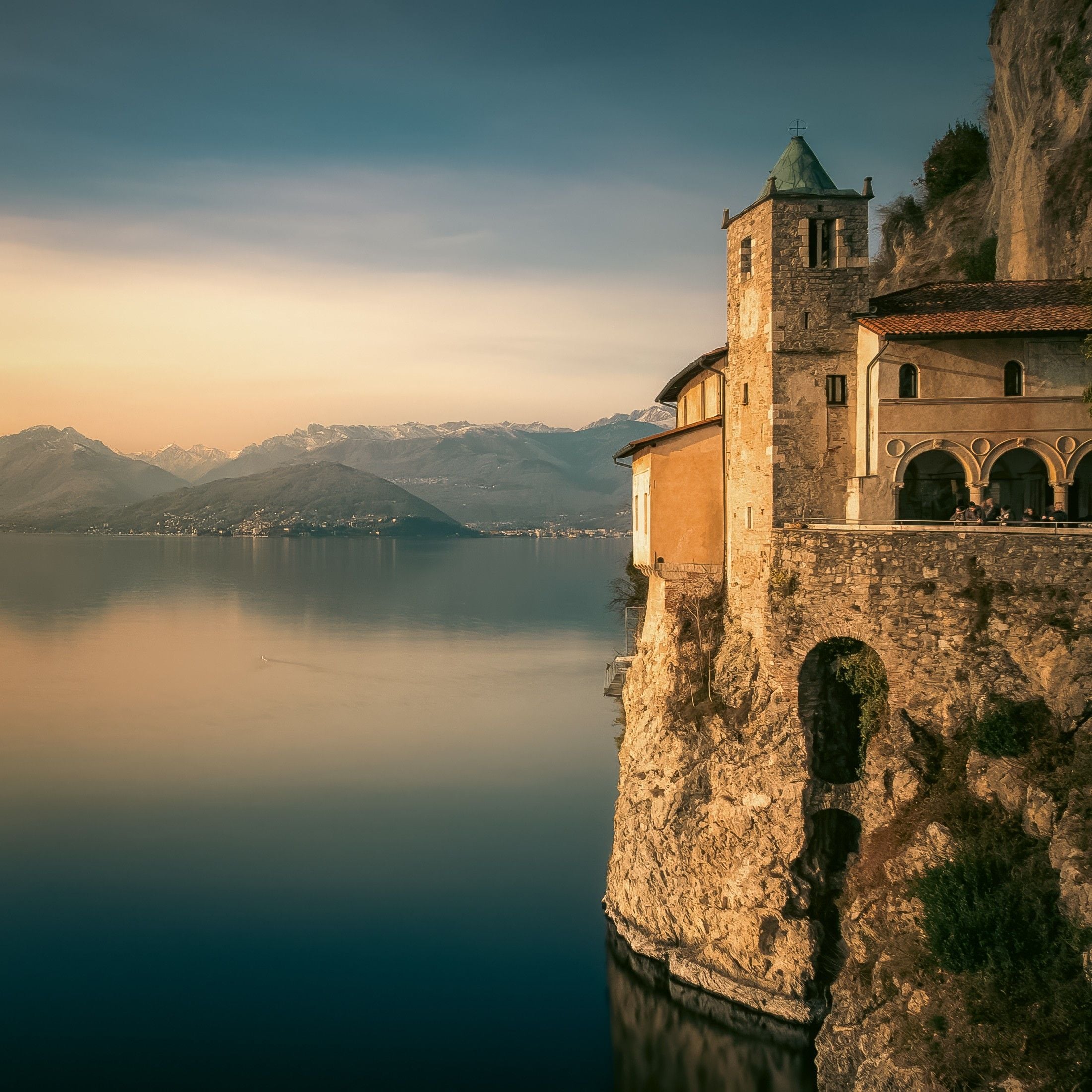 Clifftop church overlooking a tranquil lake, surrounded by mountains at sunset. Soft light enhances the serene landscape.