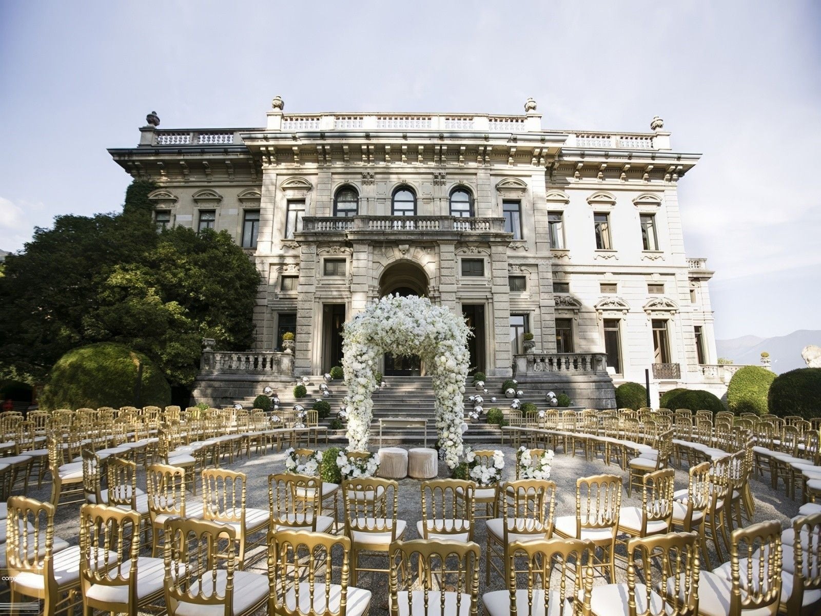 Elegant outdoor wedding setup with golden chairs and a floral arch in front of a grand mansion.