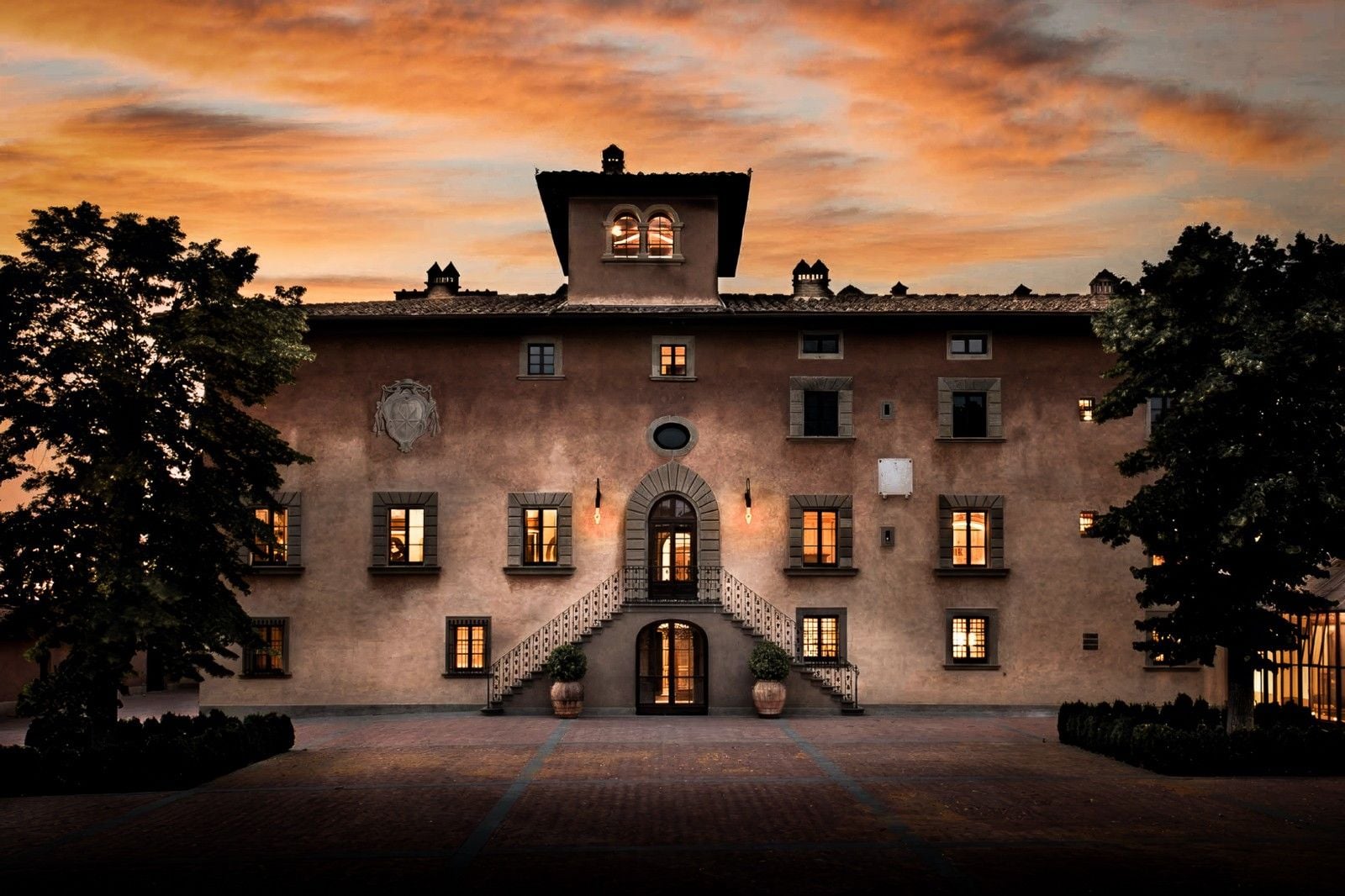 Historic villa at sunset, featuring elegant architecture, warm glowing windows, and lush greenery in the foreground.