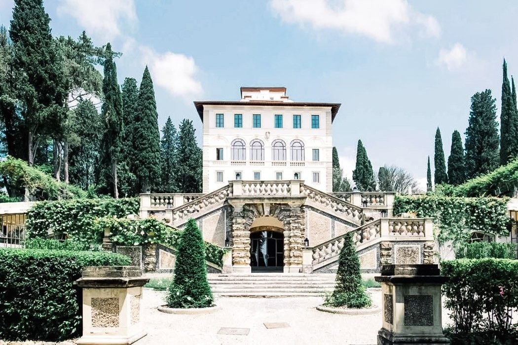 Elegant villa surrounded by lush gardens and tall cypress trees under a clear blue sky.