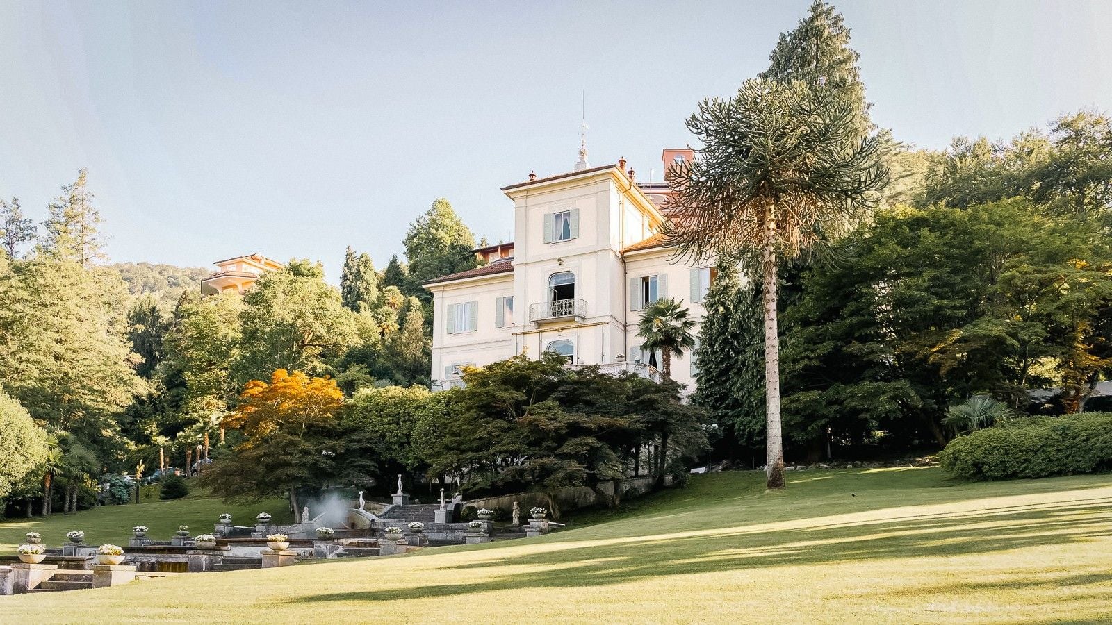 Elegant villa surrounded by lush greenery and tall trees, set against a clear blue sky.