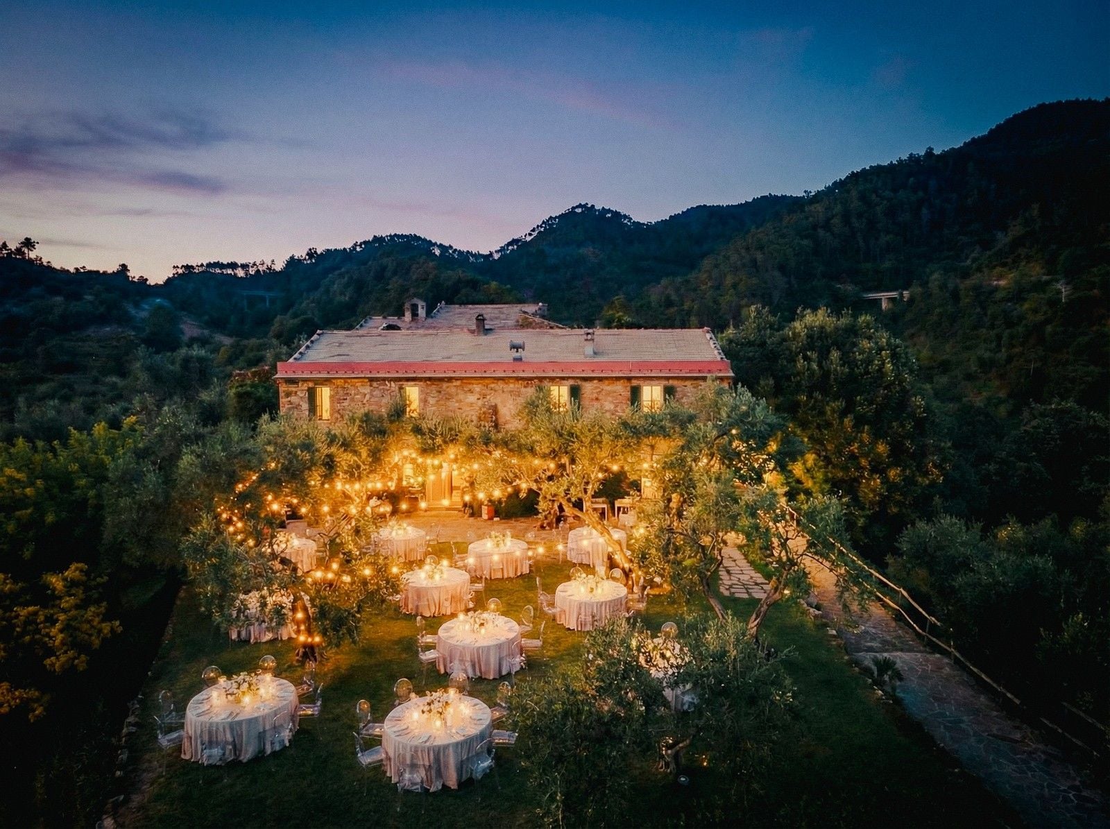 A beautifully set outdoor dining area with round tables lit by fairy lights, surrounded by trees and mountains at dusk.