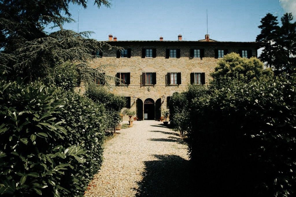 A charming stone villa surrounded by lush greenery, with a gravel pathway leading to its entrance.