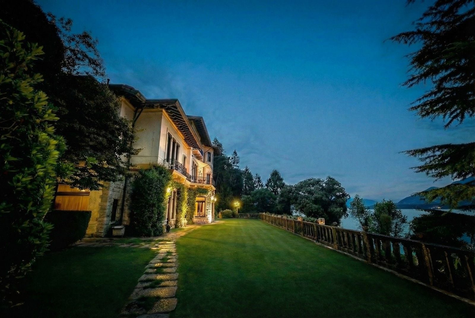 Elegant villa by a tranquil lake, illuminated softly at dusk, surrounded by lush greenery and a well-maintained garden.
