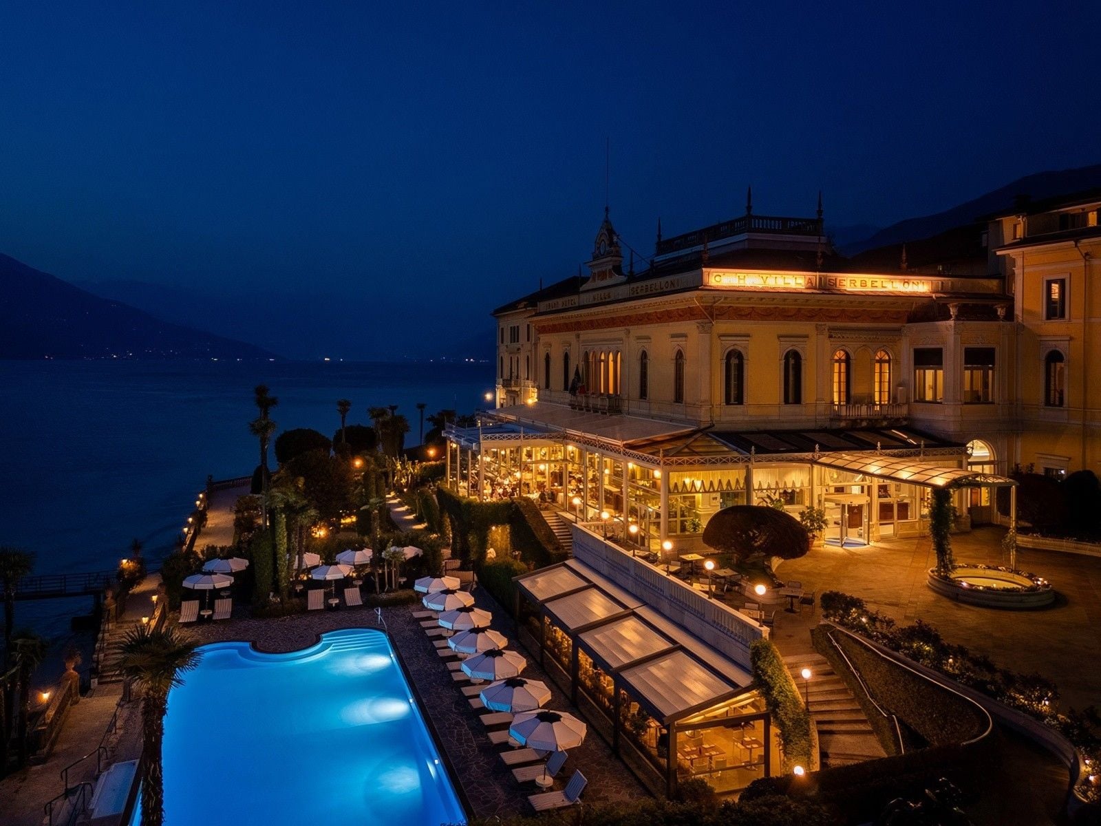 Luxurious lakeside hotel illuminated at night, featuring a sparkling pool and elegant terrace dining areas.