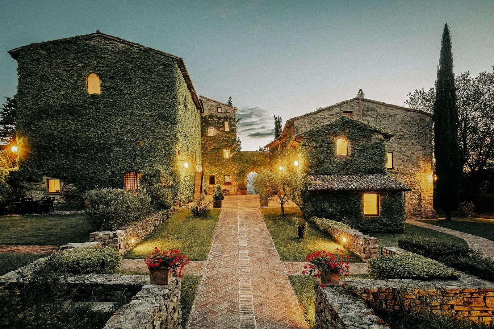 Charming stone villas covered in ivy, illuminated pathways, and lush gardens at dusk in a serene Tuscan countryside setting.