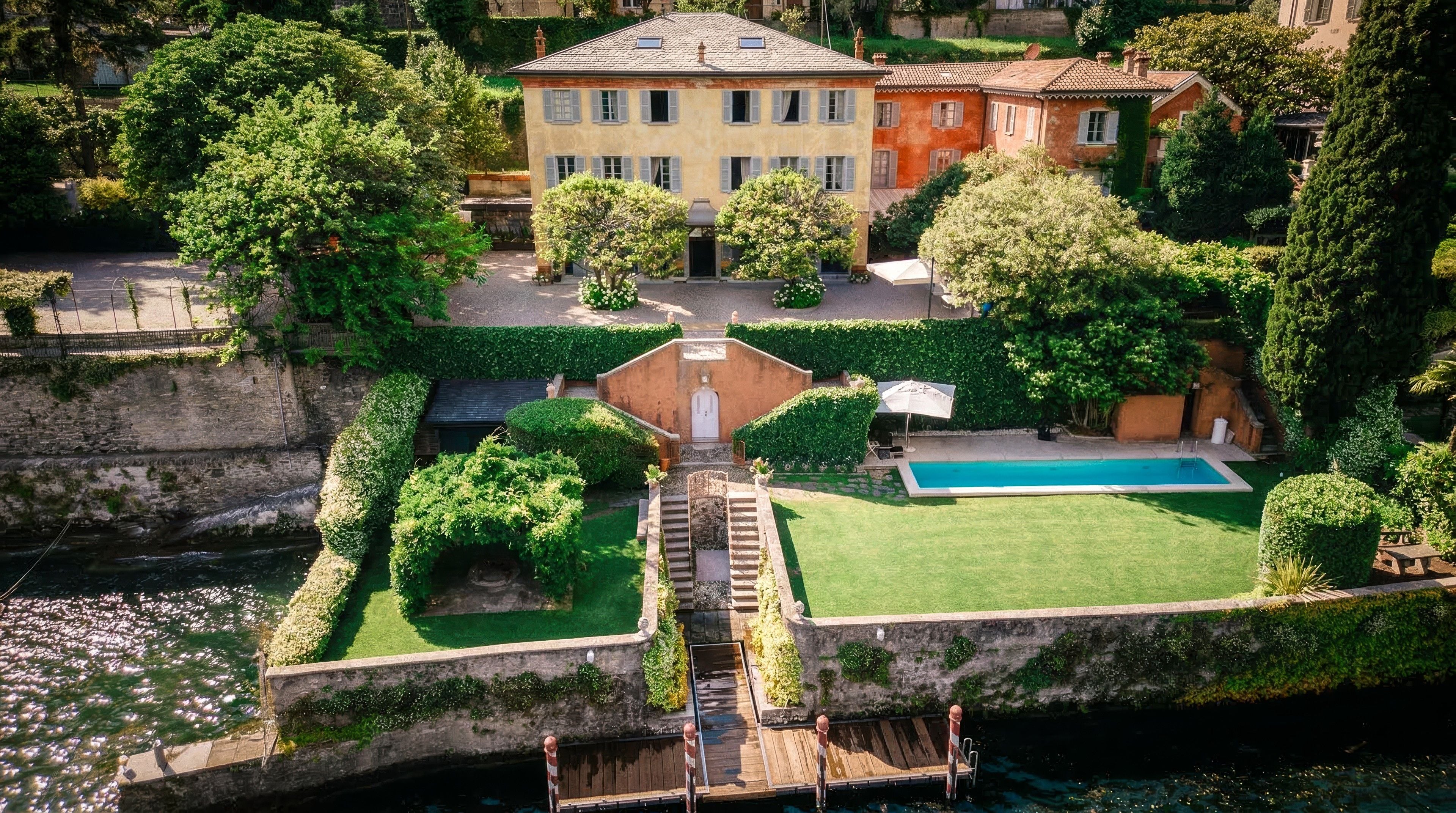 A charming villa by the water features lush gardens, a swimming pool, and vibrant greenery under a sunny sky.
