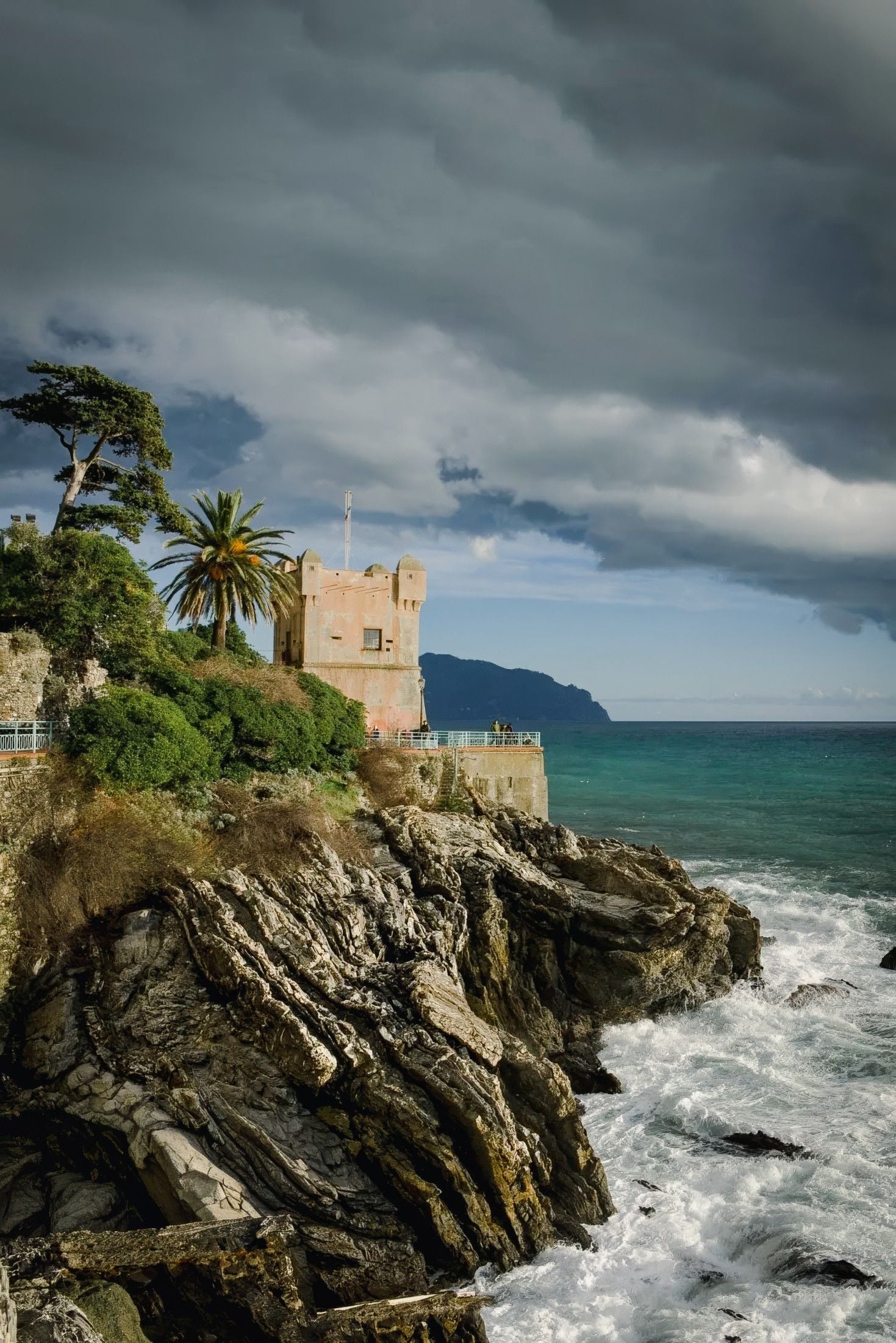 A historic tower overlooks turbulent waves crashing against rocky cliffs under a moody, cloud-filled sky.