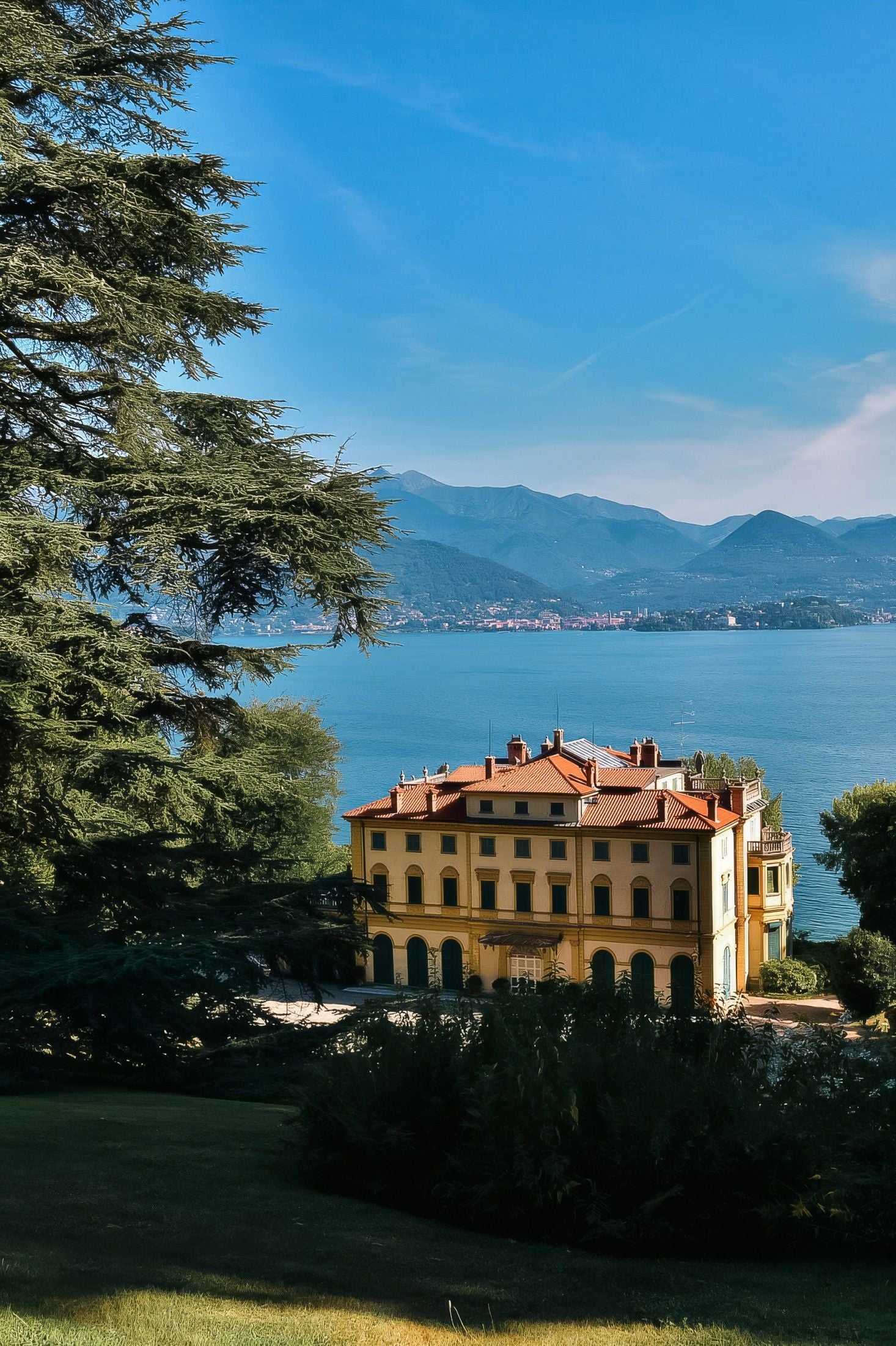 A yellow villa with a red-tiled roof overlooks a serene lake surrounded by mountains and lush greenery.