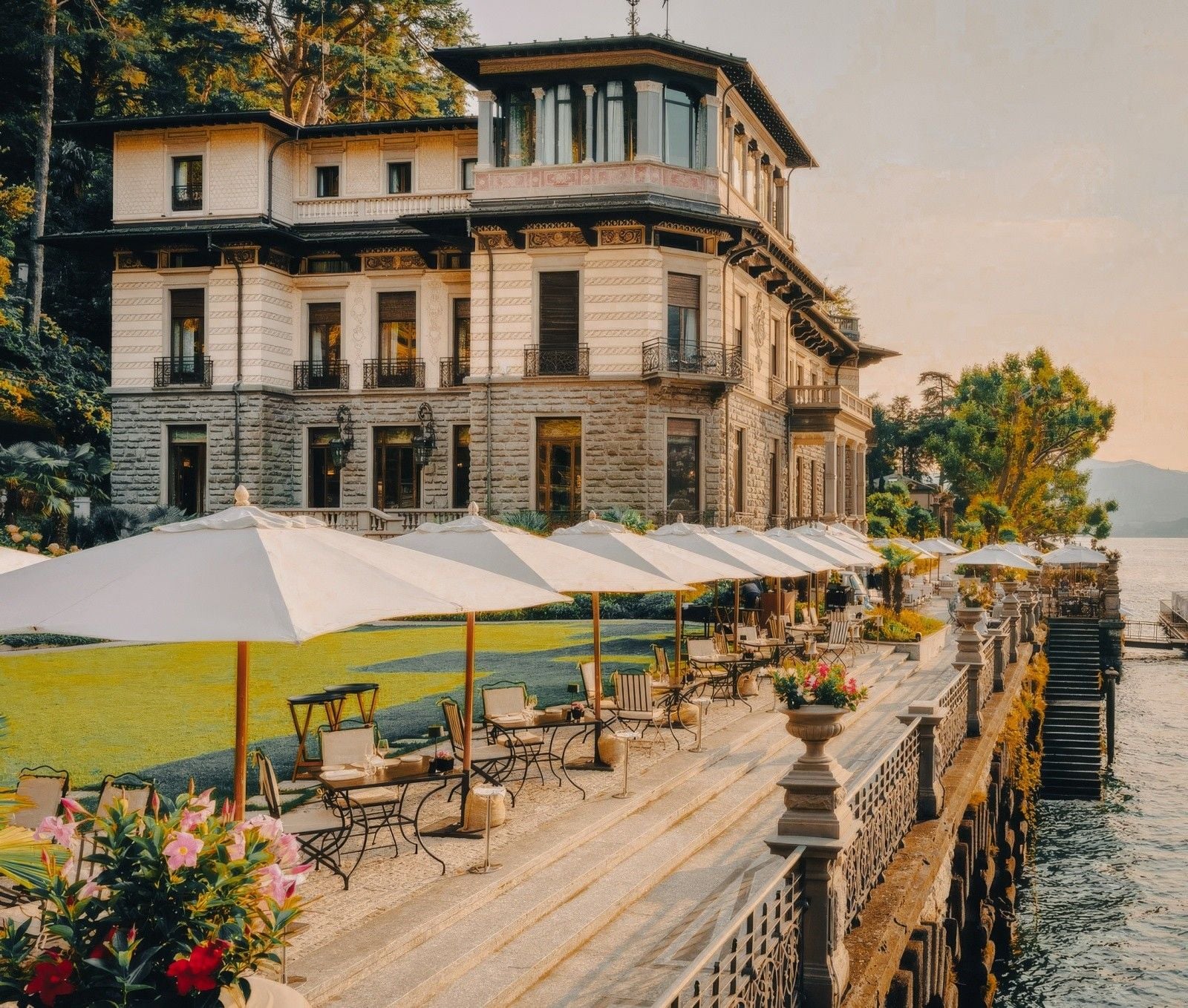 Elegant waterfront villa with outdoor seating and parasols, surrounded by lush greenery and a tranquil lake.