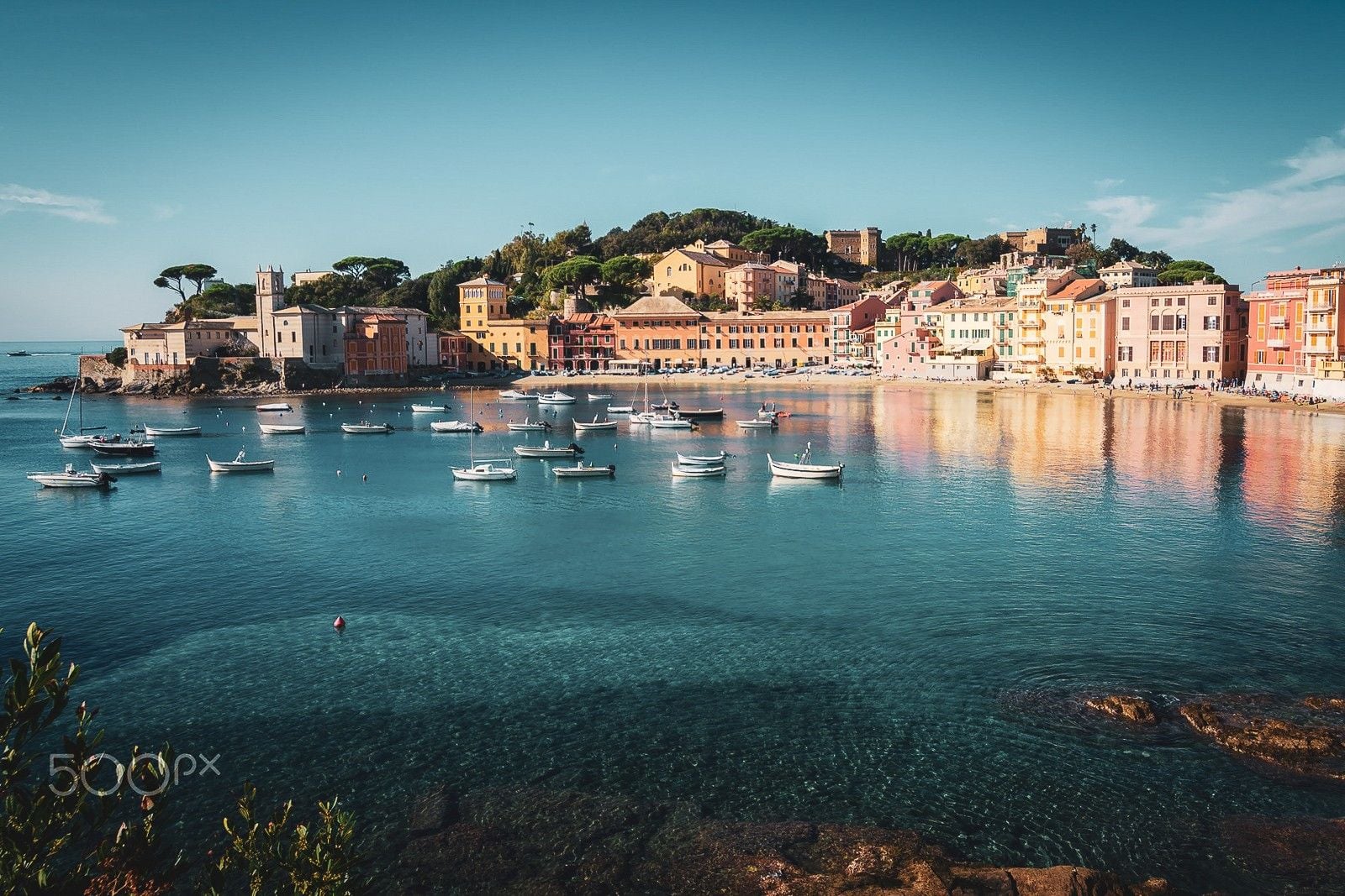 Colorful buildings line the harbor of a serene coastal village, with boats gently bobbing in the clear blue water.
