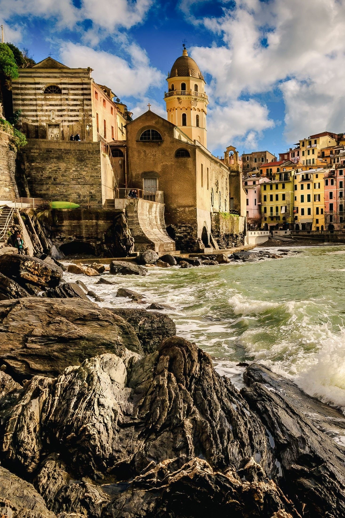 Colorful buildings line the rocky shoreline, with a historic church perched above the churning sea in a coastal village.