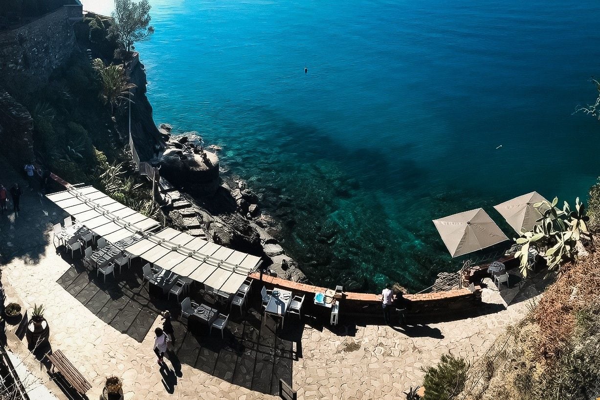 Coastal restaurant overlooking clear blue waters, featuring outdoor seating and rocky shoreline.